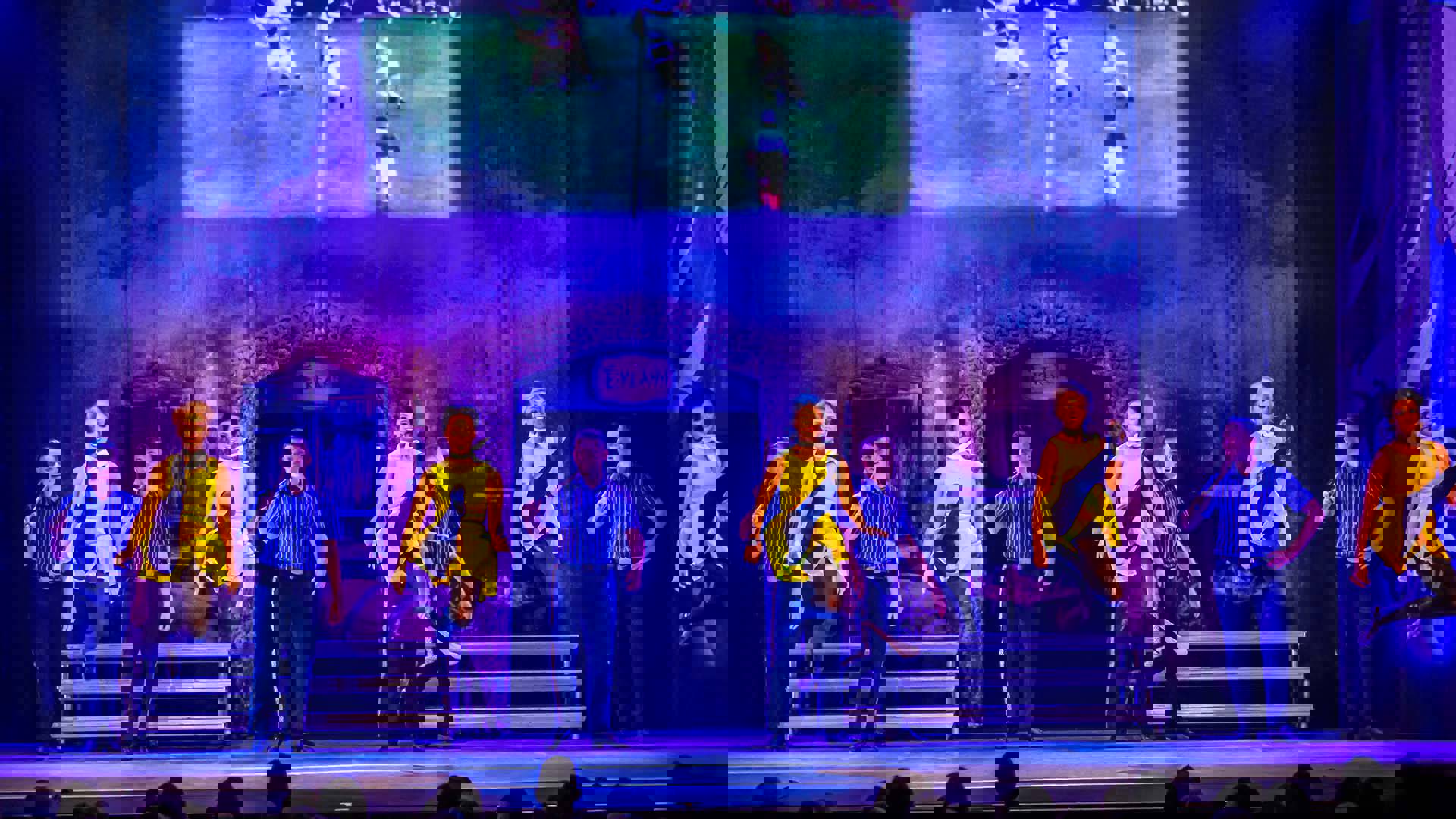 A line of women in yellow dresses leap into the air. A line of men in blue shirts stand behind them.