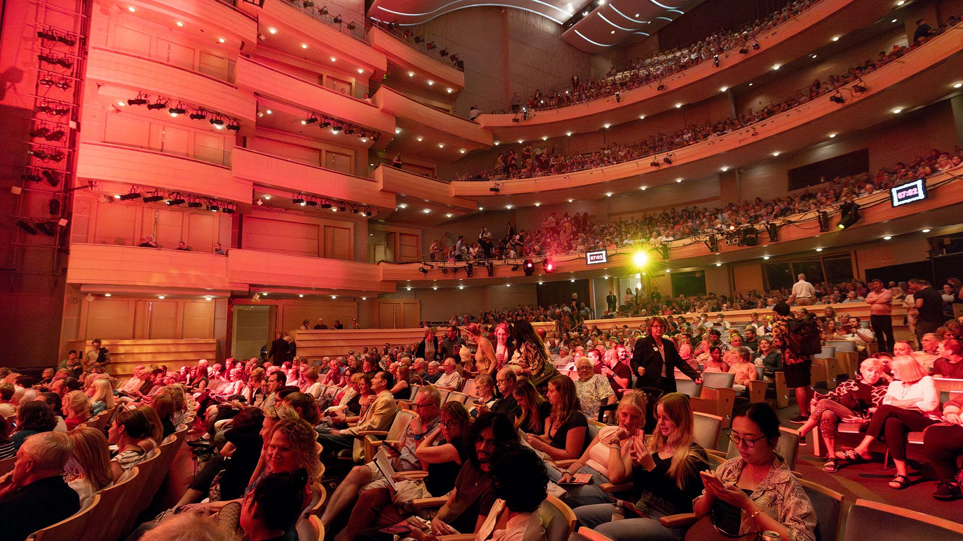 A packed audience in Overture Hall. The orchestra level is filled with seated attendees, the house is lit a soft red with views up to the balcony.