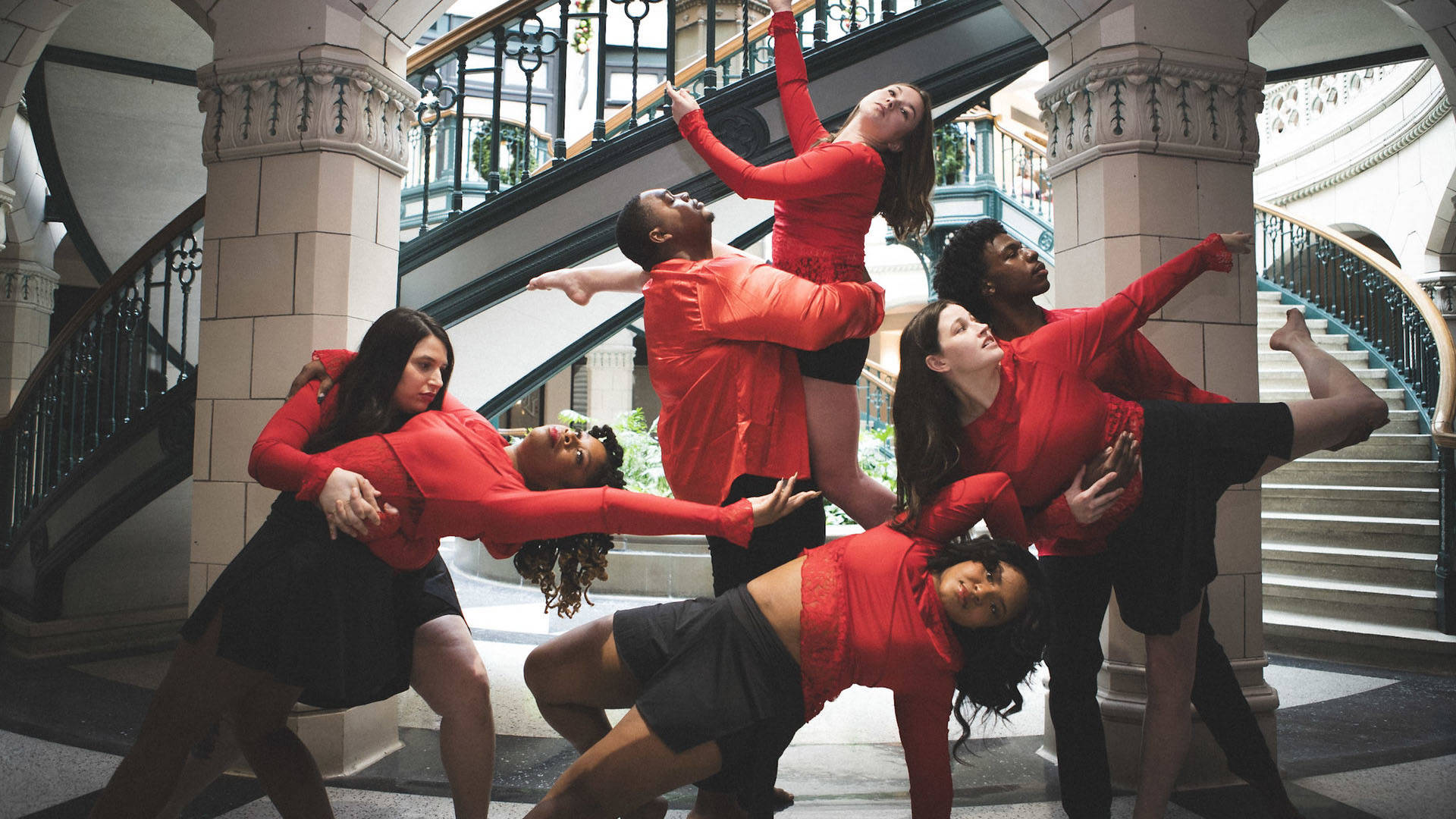 Seven dancers with a diversity of dark, medium and light complexions wearing red longsleeve shirts and black shorts are posing with each other in stretching/reaching movements in front of a white columned spiral staircase.