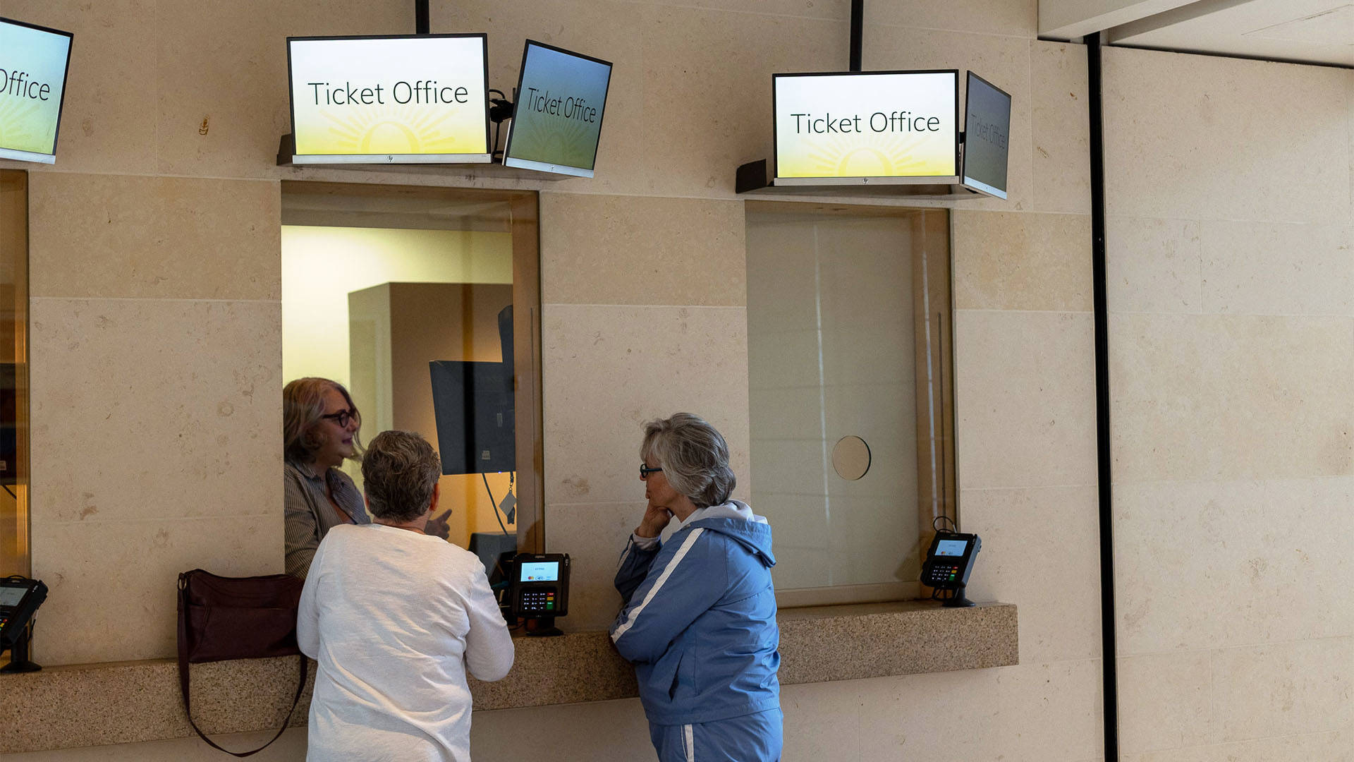 Two older caucasian women standing in front of Overture's Ticket Office being helped by an employee in a window.