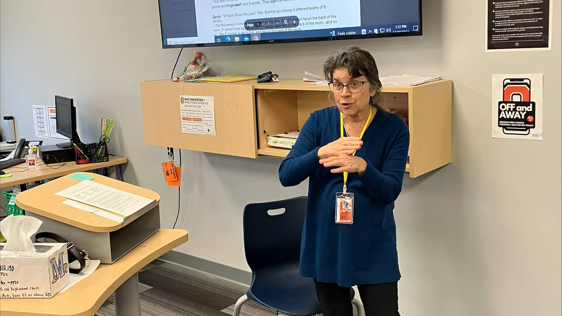 Michelle Guyette communicating in ASL in a classroom. She is a middle aged woman with greyish brown hair, light complexion, and glasses in a blue top.