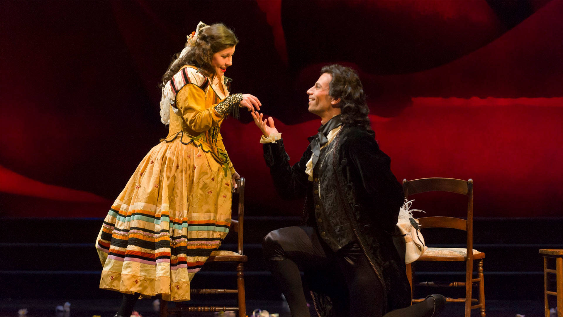 Performers in the opera "Don Giovanni" performing on a stage. A man in fine dark clothing with dark hair kneels and holds the hand of a woman in a fine golden dress with long brown hair.