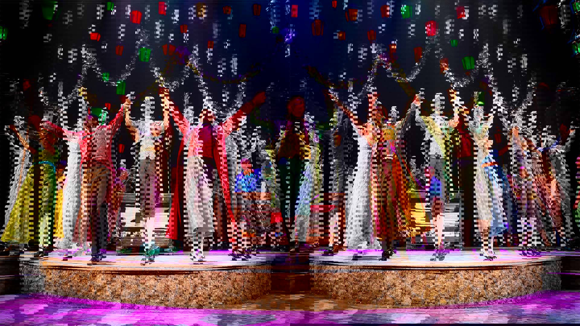 The cast of "A Christmas Carol" holding hands in a row with arms raised, smiling. They are in period style, colorful costumes from 19th century England. Green, red and yellow lit lamps hang above.