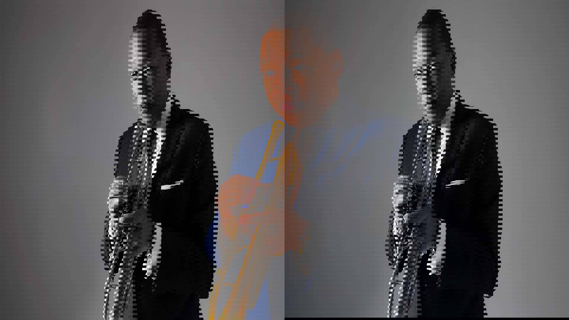 Wynton Marsalis standing in front of a grey backdrop. He has a cool blue suit, is holding a trumpet in playing position and looks confidently towards the viewer. He has short brown hair and dark eyes.