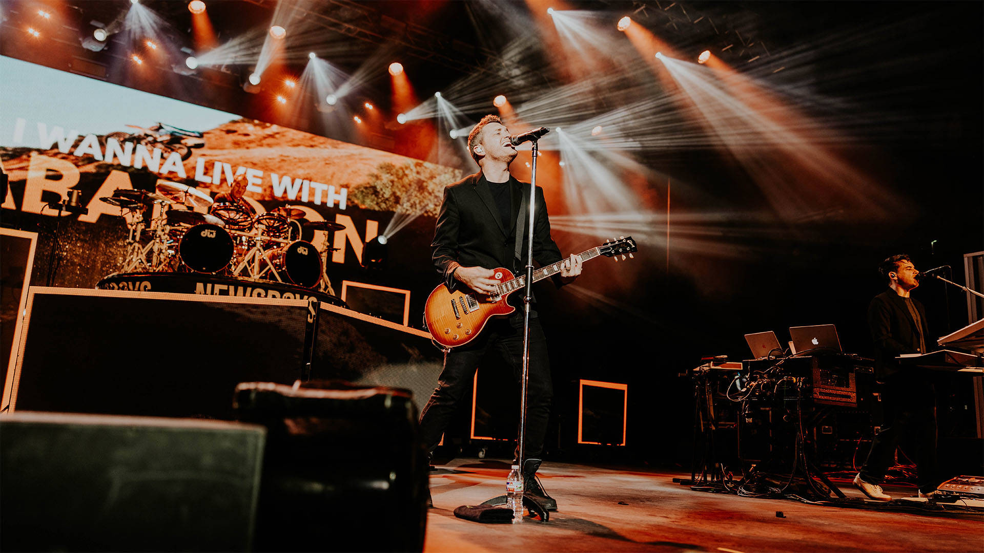 A man playing guitar and singing into a microphone on a stage. He has light complexion and short blond hair.