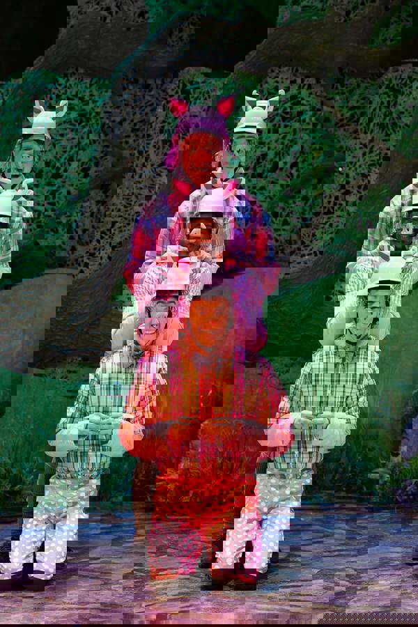 Three men, two caucasian and one black, smiling and wearing pink outfits and pink construction hats with pig ears attached. They are dressed as the three pigs and standing in a line.