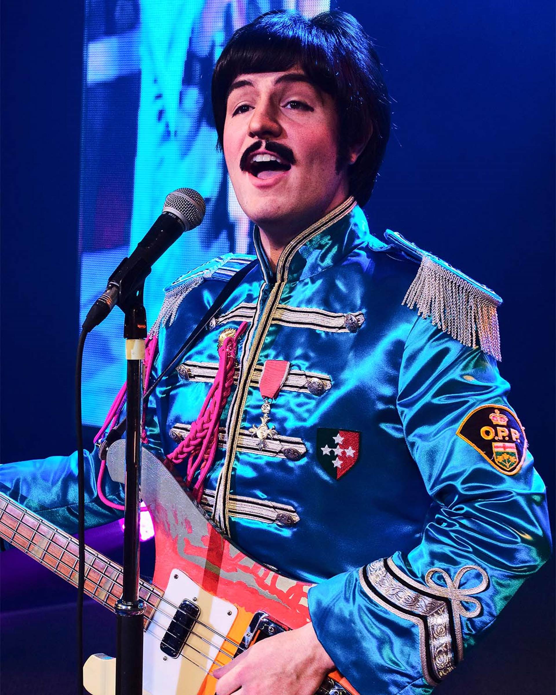 A Paul McCartney impersonator wearing a blue Seargent Peppers outfit and holding a guitar. He has dark hair and a moustache