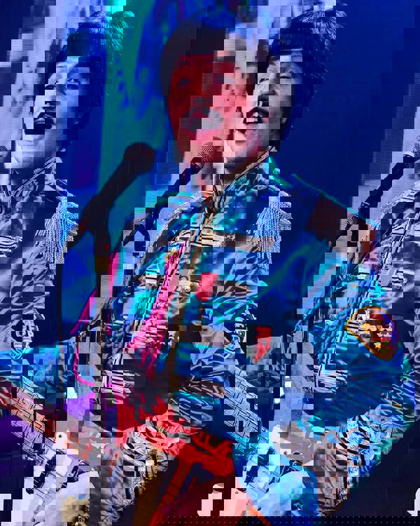 A Paul McCartney impersonator wearing a blue Seargent Peppers outfit and holding a guitar. He has dark hair and a moustache