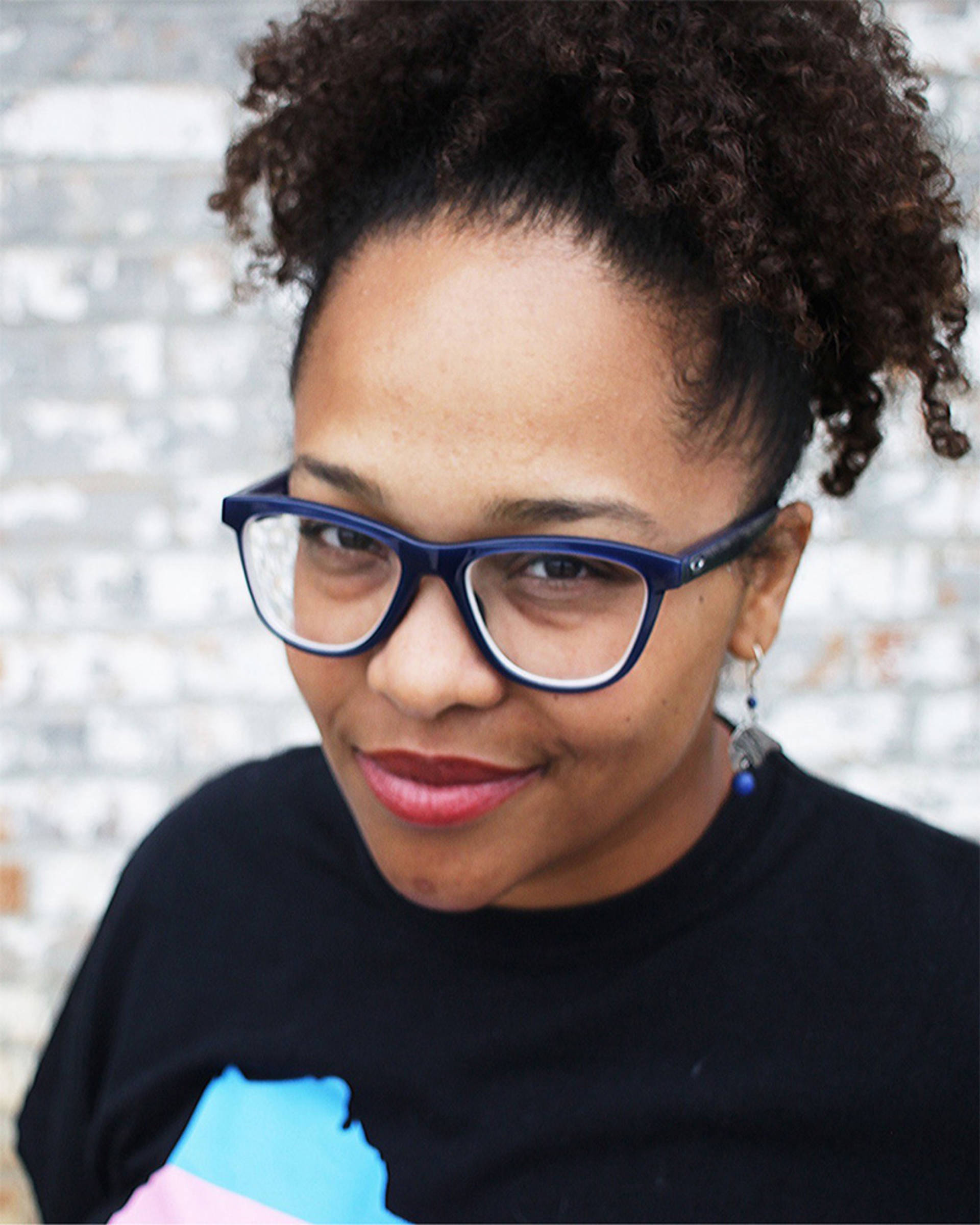 Headshot of a young african american woman with curly dark hair worn up, a black tshirt, and blue glasses.