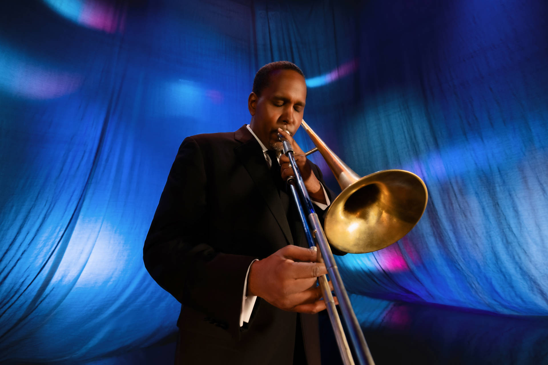 A man with dark complexion and black hair plays a trombone in front of a blue stage curtain.
