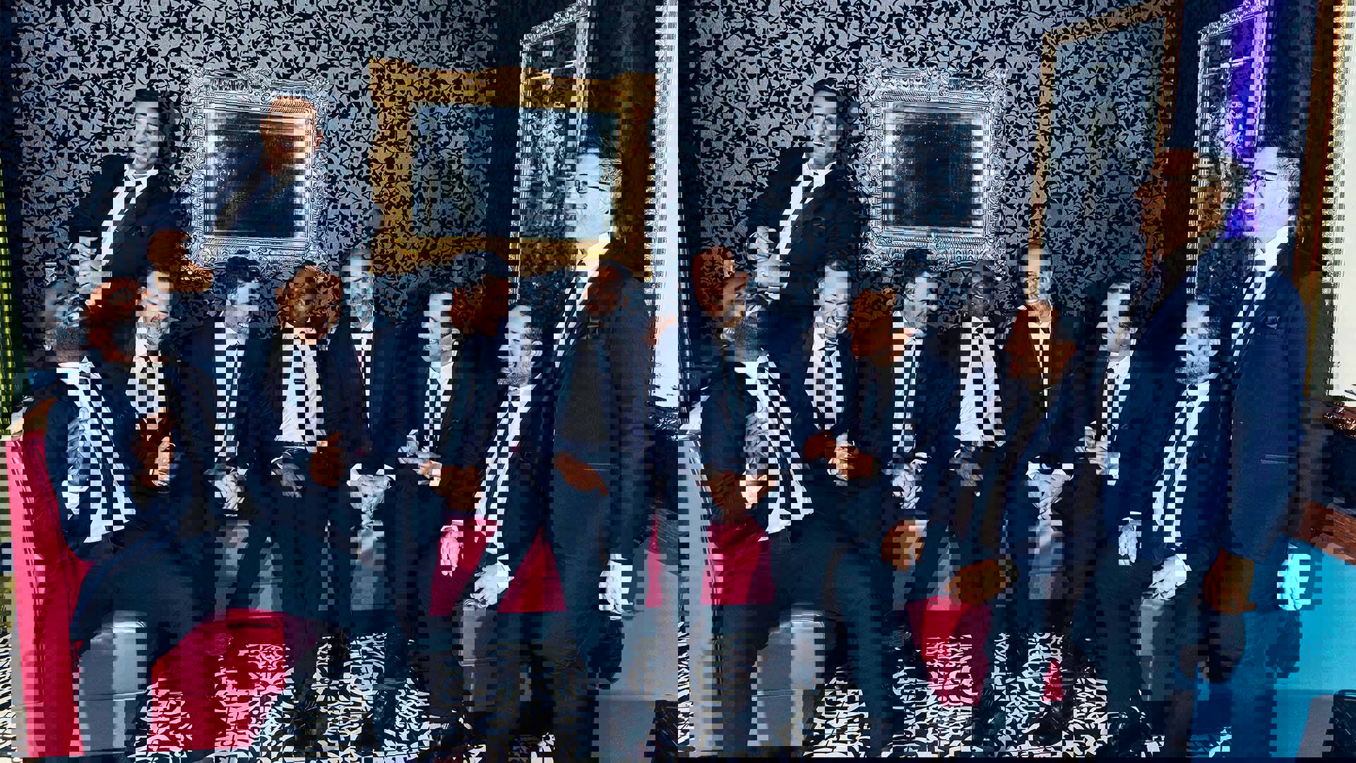 Nine men wearing blue suits sitting on a pink couch. They are laughing and looking like they're having a grand time.