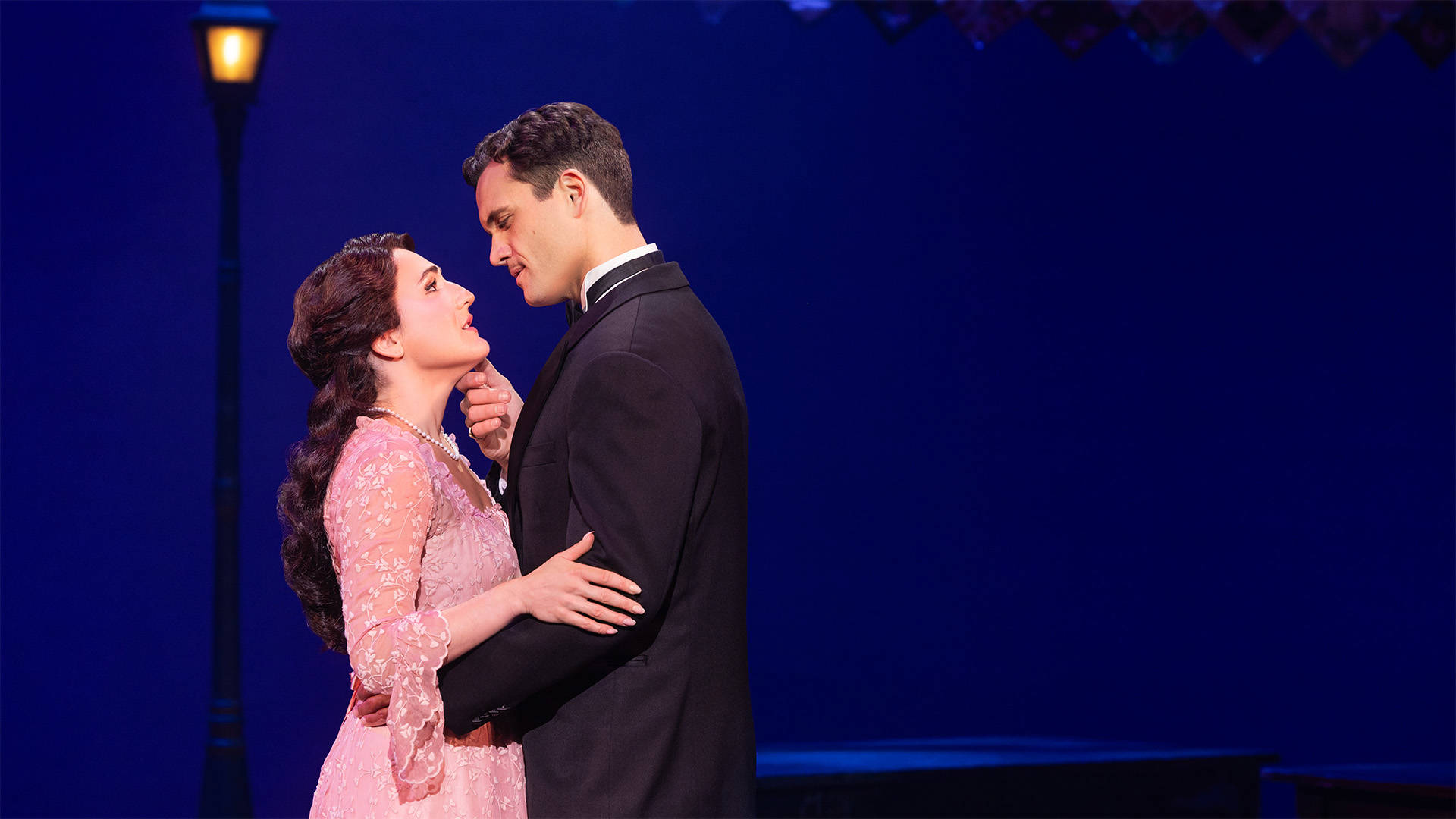 A man and a woman embrace eachother against a dark blue backgorund. The woman has light complexion and long brown hair. The man has light complexion, a moustache and brown hair.