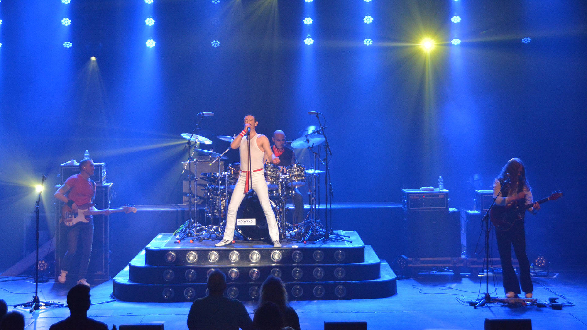 A Queen cover band featuring a singer in a white outfit impersonating Freddie Mercury. The stage is cast in blue lights