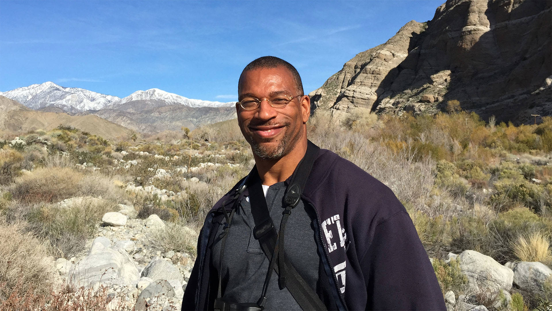 Christian Cooper smiling in a desert landscape with mountains in the background. He is a middle aged man with dark complexion, buzzed hair and short goatee, with glasses and dark clothing.