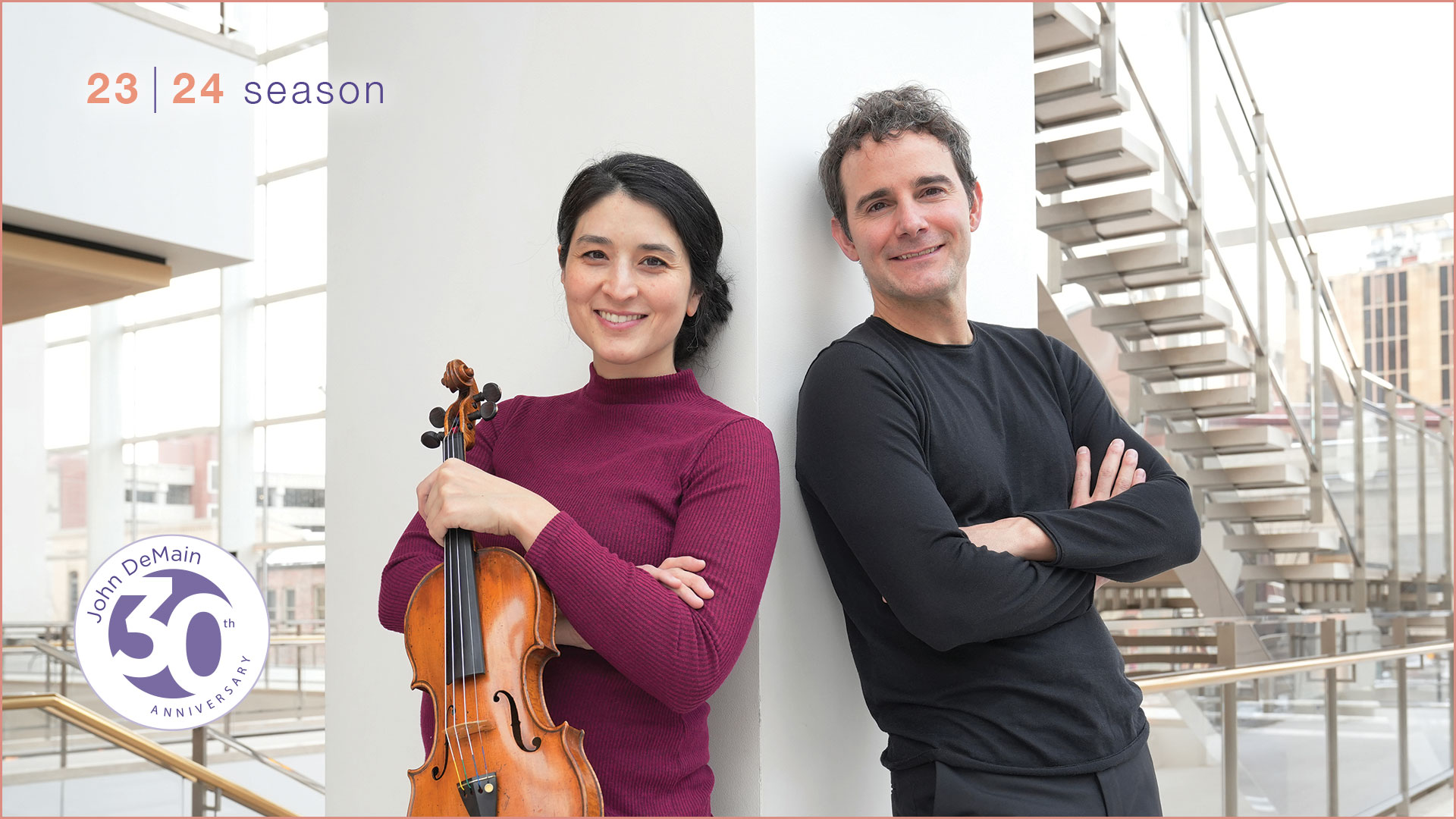 A young man and woman lean against a white pilar in Overture Hall lobby. The woman has dark hair and is holding a violin. The man has a black top on with curly graying hair.