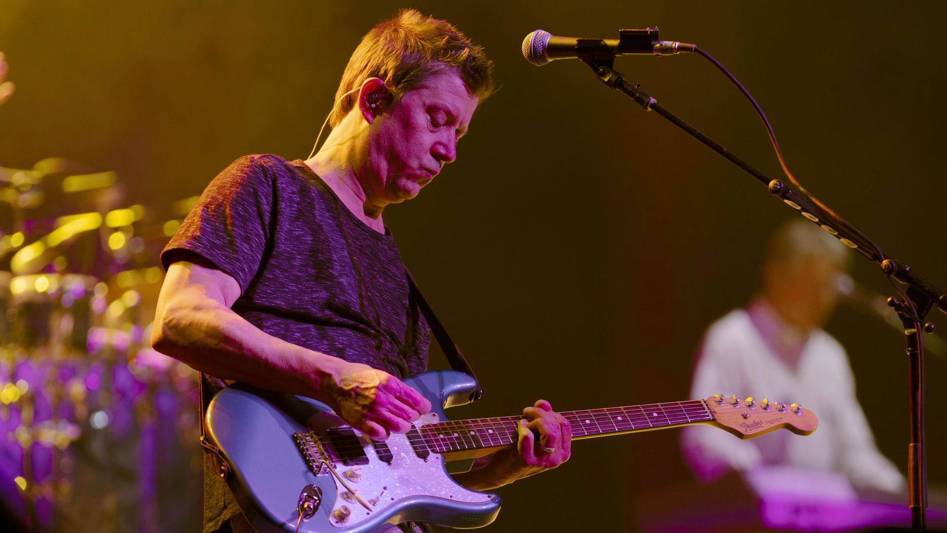 A white man standing in front of a vocal mic playing a blue guitar.