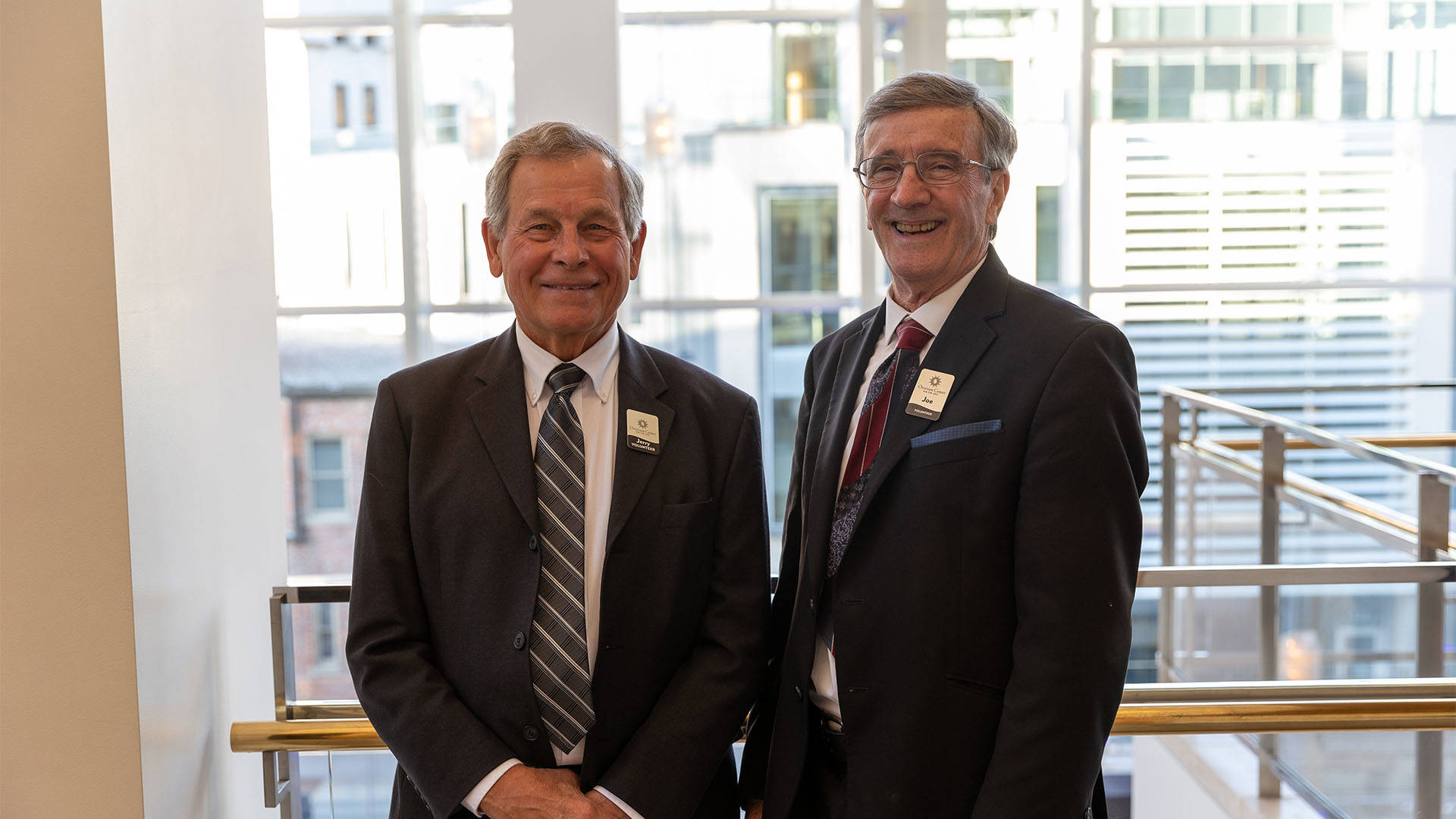 Two older caucasian men in dark suits pose for a photo together.