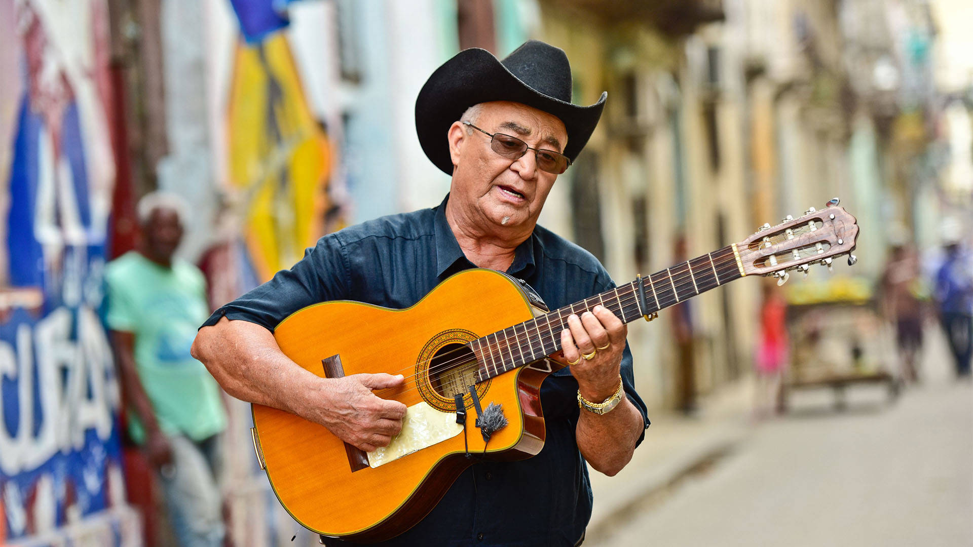 Eliades Ochoa, a Cuban man with medium skin tone and short white hair. He is wearing tinted glasses, a black cowboy hat and a black dress shirt while playing the guitar in a street.