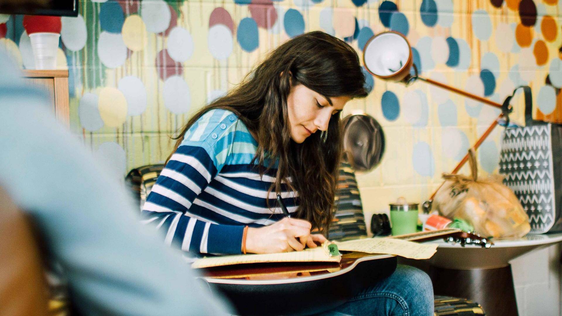 A young woman with medium complexion and long dark hair sits and writes on a notepad that is resting on an acoustic guitar on her lap. A colorful painting brick wall is behind her.
