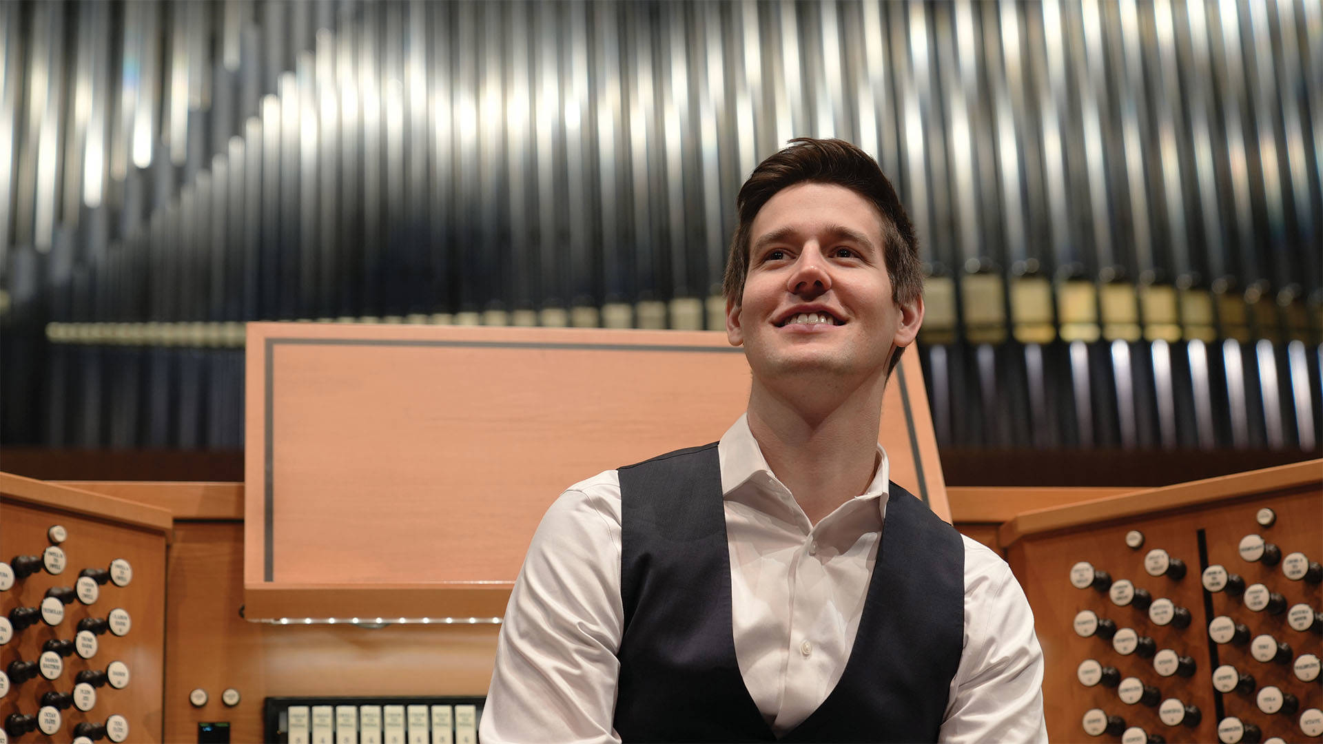 Greg Zelek, a young man with light complexion, short styled brown hair, a white button-up shirt and black vest, smiling in front of an organ console and large metalic pipes