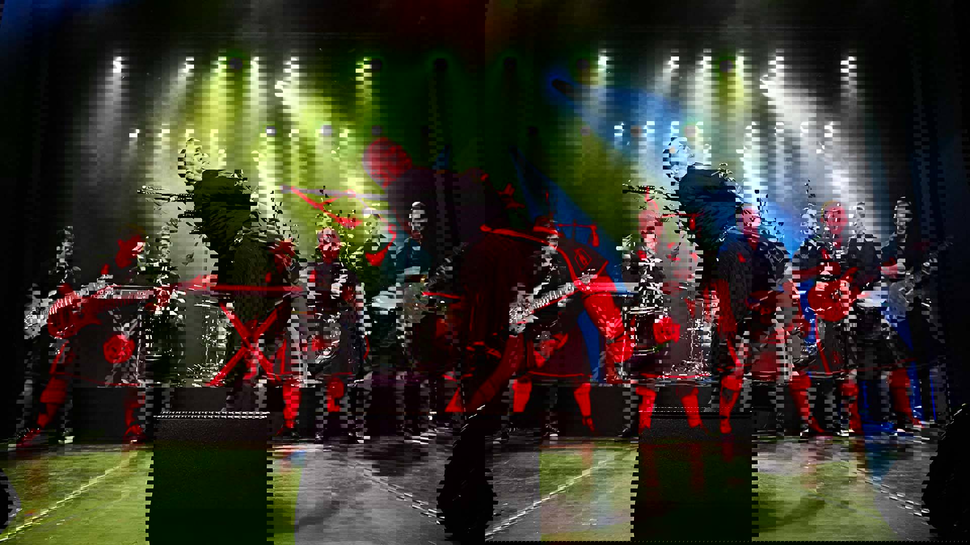 Dramatic image of the Red Hot Chilli Pipers performing on a stage under green and blue lights. they are wearing matching black outfits and kilts with red highlights and long red socks. Instruments include bagpipes, guitars, a marching snare drum and a djembe.
