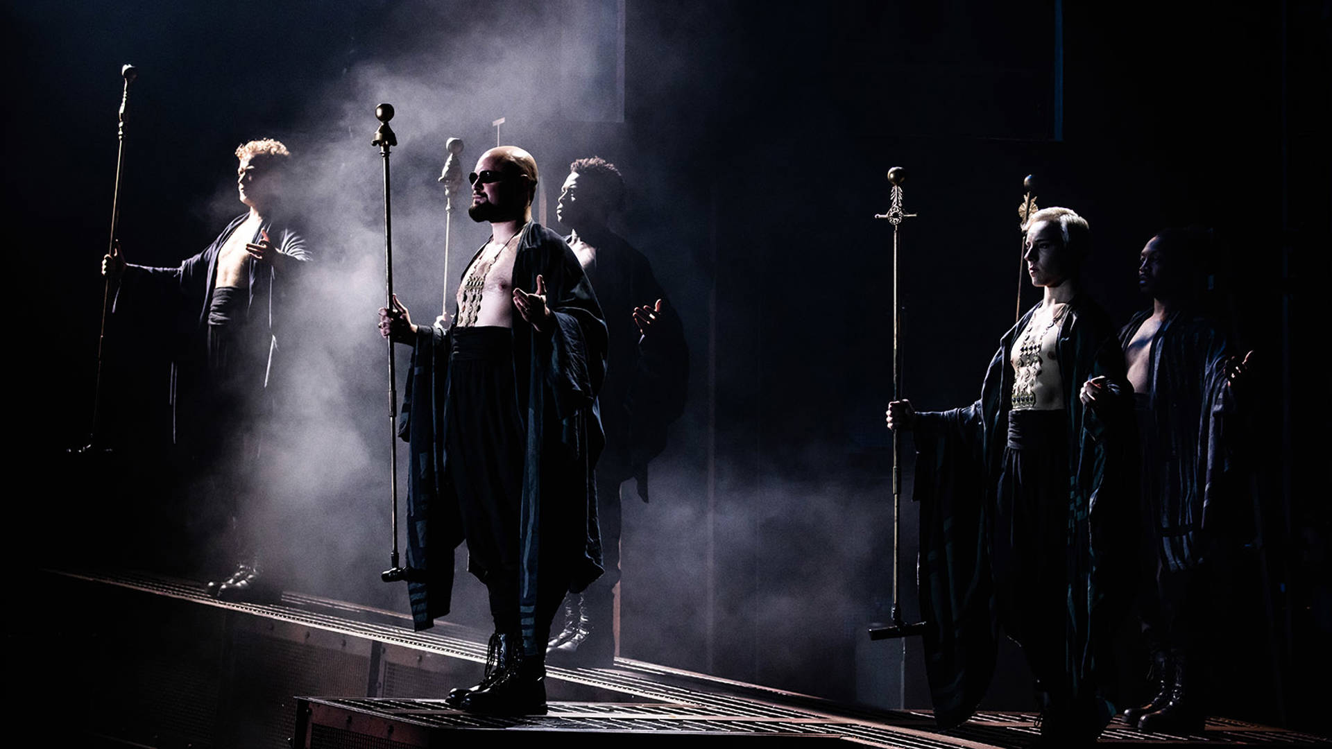 Five cast members stand facing the side holding long metal staffs. They are shirtless with black bottoms and shawls.
