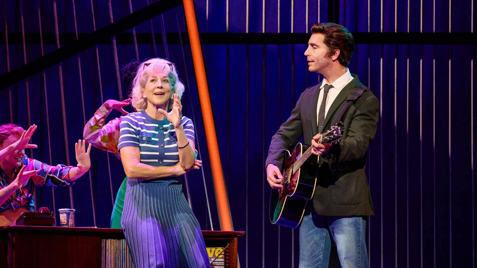 A man with light complexion and greased brown hair, in a dark suit with tie playing an acoustic guitar and facing a woman with greyish blonde hair in a blue outfit, holding her left hand in the air.
