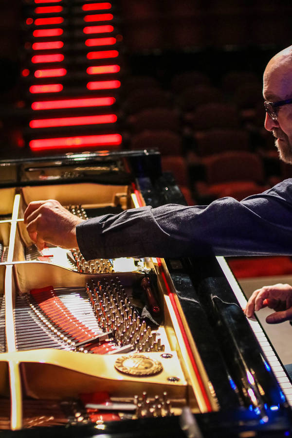 George Winston playing a piano with one hand reaching towards the strings. He is a caucasian man with a bald head, grey beard, and glasses