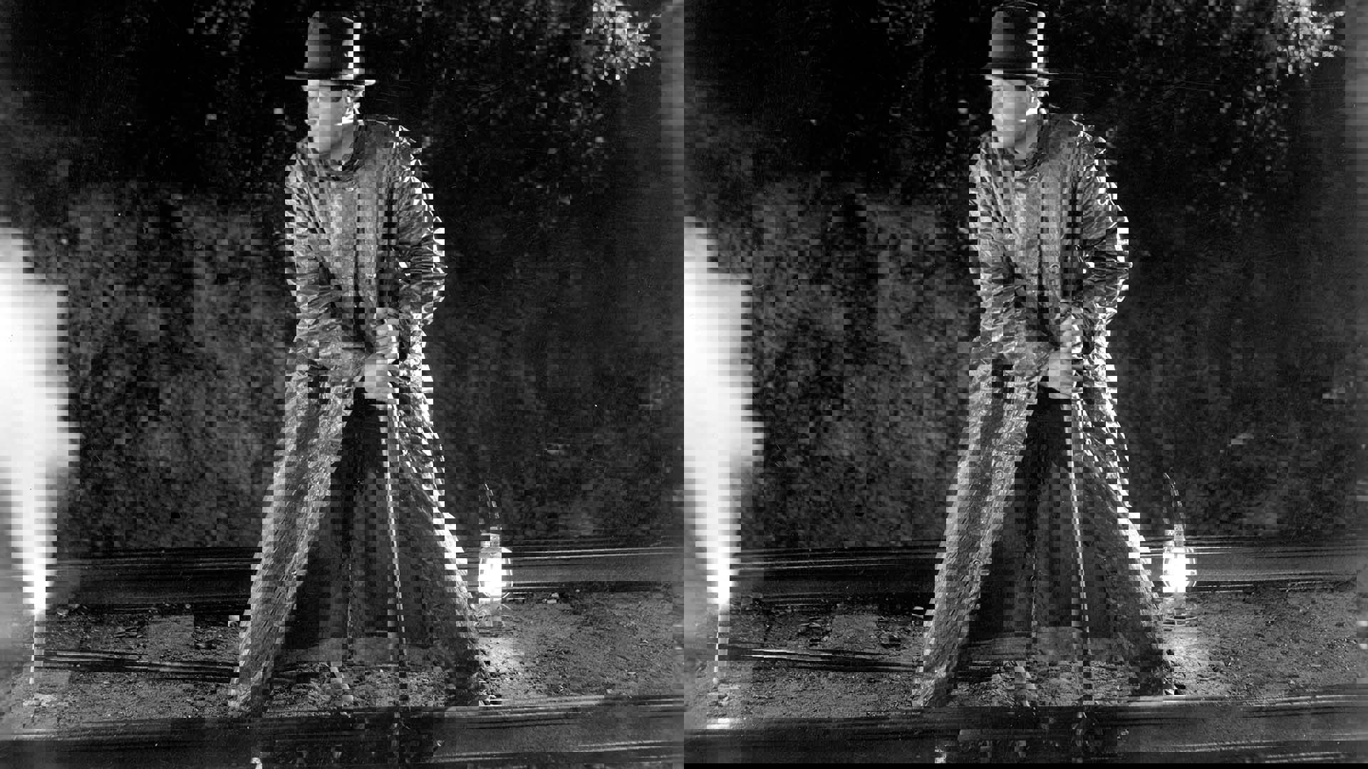 Dave Taylor (played by Rockliffe Fellowes), a light complexion man in a fedora hat and trenchcoat, uses a long wrench to loosen a railroad tie while a train approaches out of the frame in a scene from "The Signal Tower (1924)."