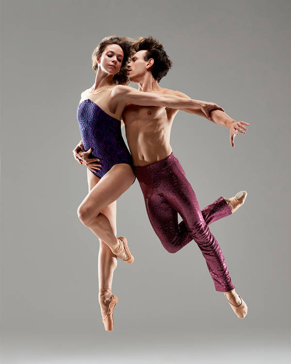Two dancers suspended in midair against a grey background. The woman on the left has light complexion and a blue outfit. The man on the right has light complexion and burgundy pants.