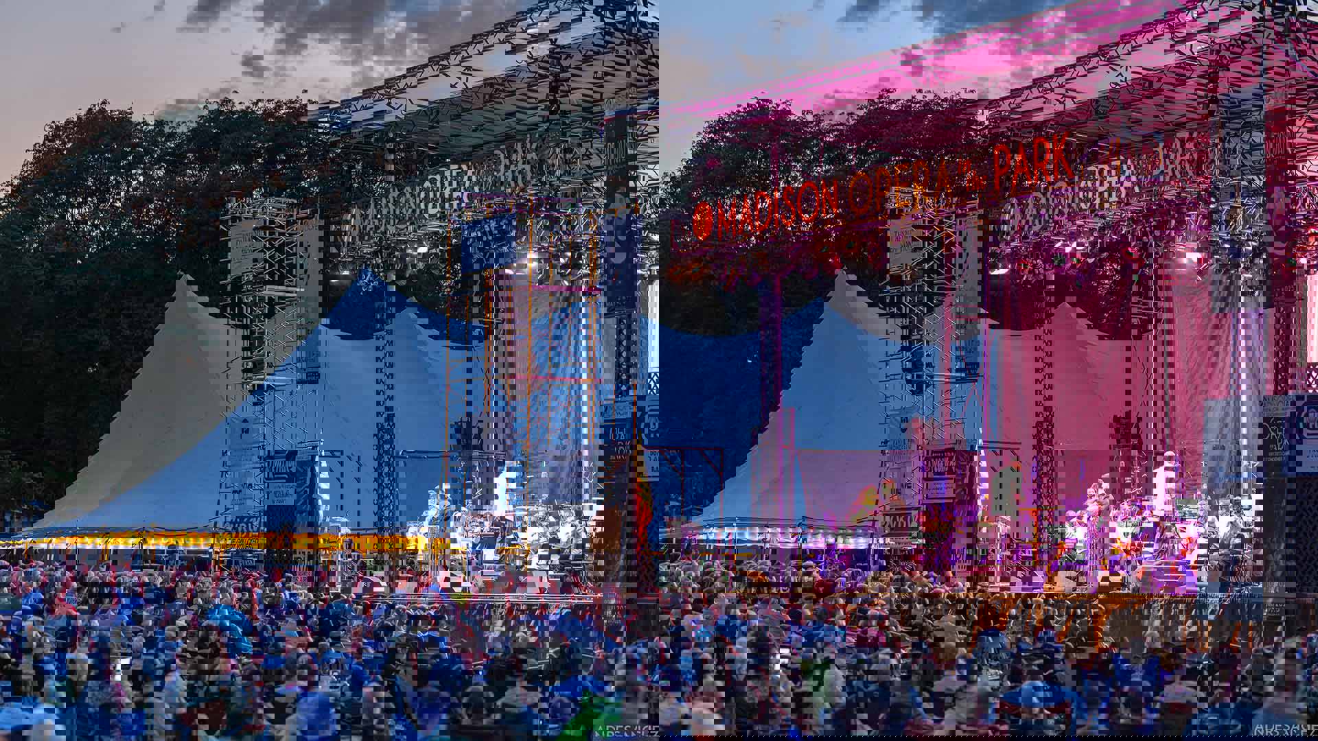 A large outdoor stage next to a large white tent with a seated audience of hundreds at dusk with trees behind. The stage is lit purple with an orchestra, conductor and two singers performing. It reads "Madison Opera in the Park" at the top.