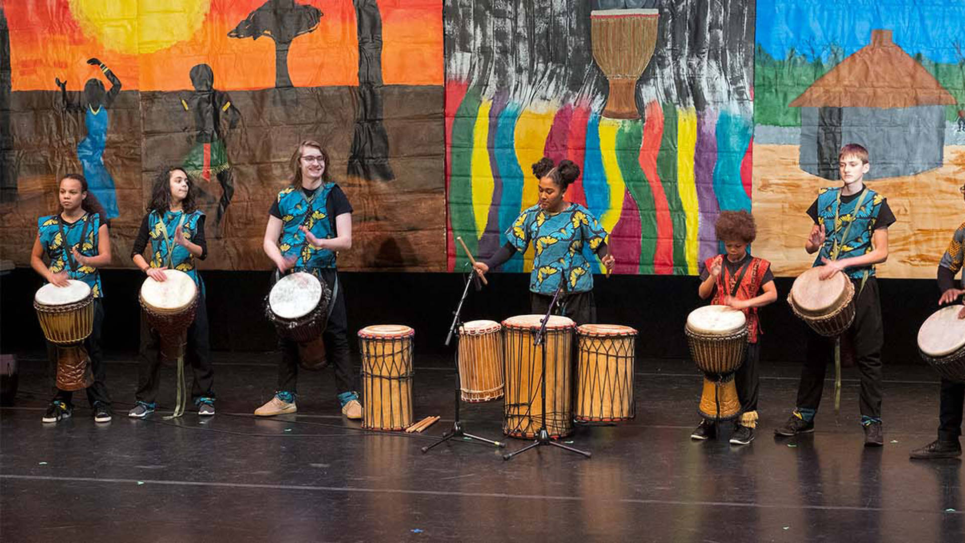 Six young performers wearing blue patterned vests each hand-drumming on stage.