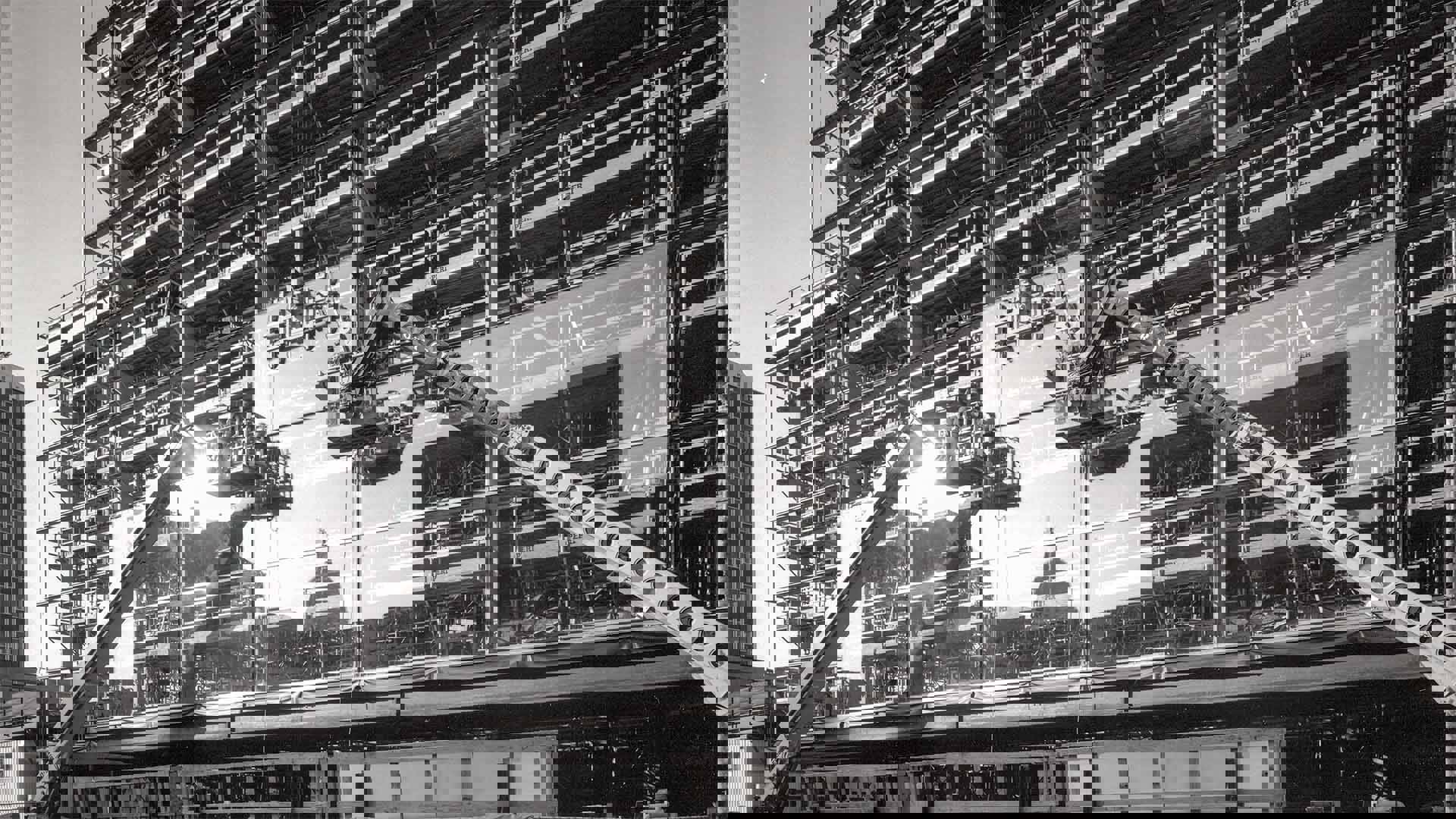 Two cranes with hard hat-wearing workers in caged platforms adding large glass windows to the steel bar exterior of Overture Center under construction. The capitol building is shown in the reflection of the glass panels already in place.  Black and white image.
