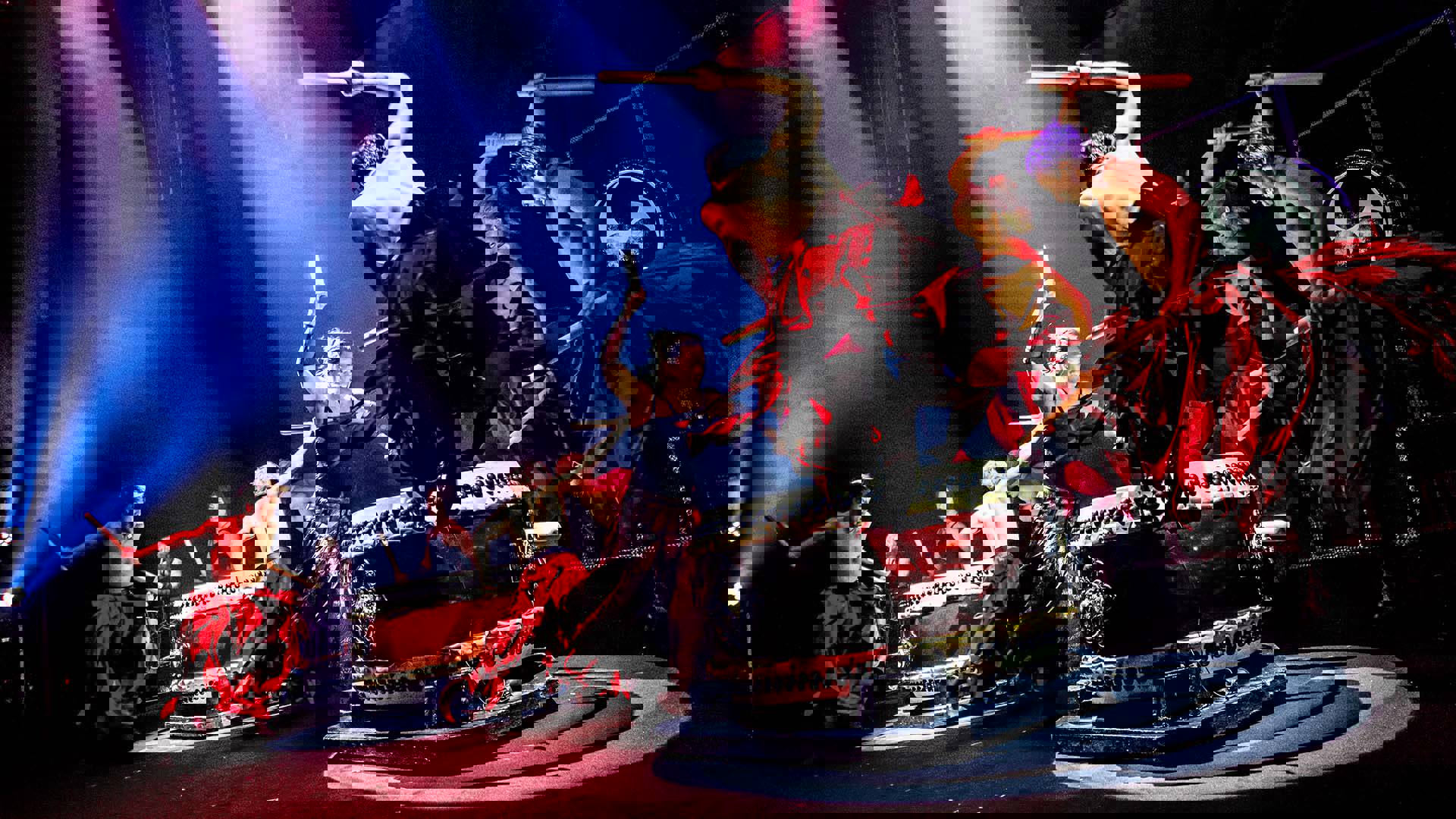 Drum Tao performers, 8 shirtless men with light to medium complexion wearing long red and black skirts, enthusiastically banging on large drums on stage with wooden sticks.