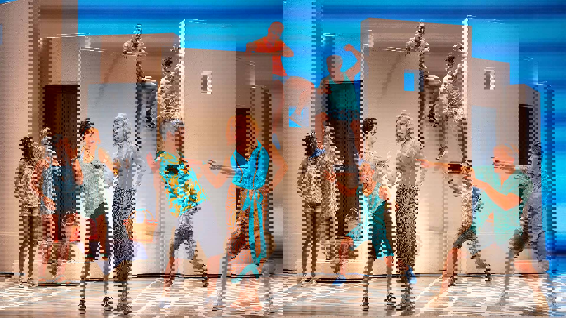 The Mamma Mia ensemble in summertime outfits in various poses against a creme building like structure and blue background.