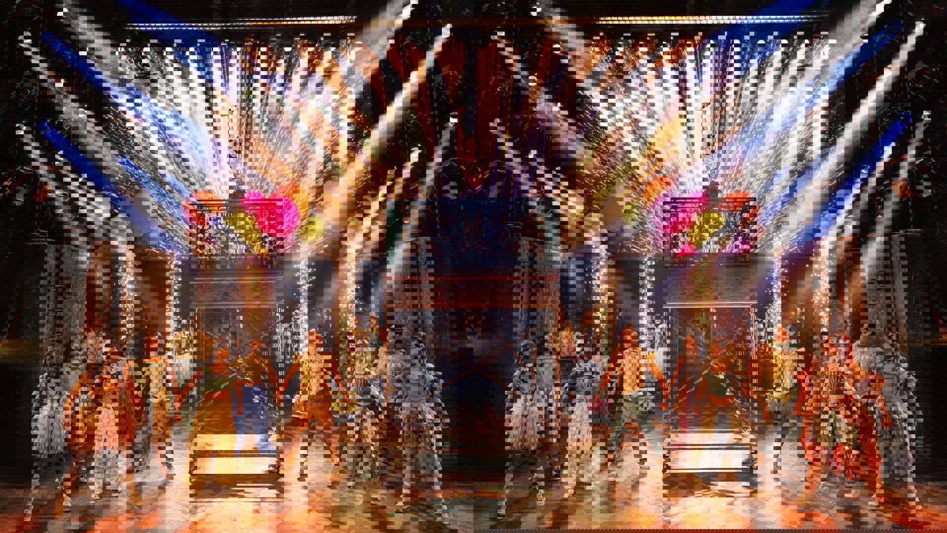 A woman stands on an ivy-covered balcony lifted high in the air. She is wearing a pink and gold crystal bodysuit, smiling with a gold microphone extended upwards in her right hand. Dancers stand on stage on either side smiling, looking up at her.