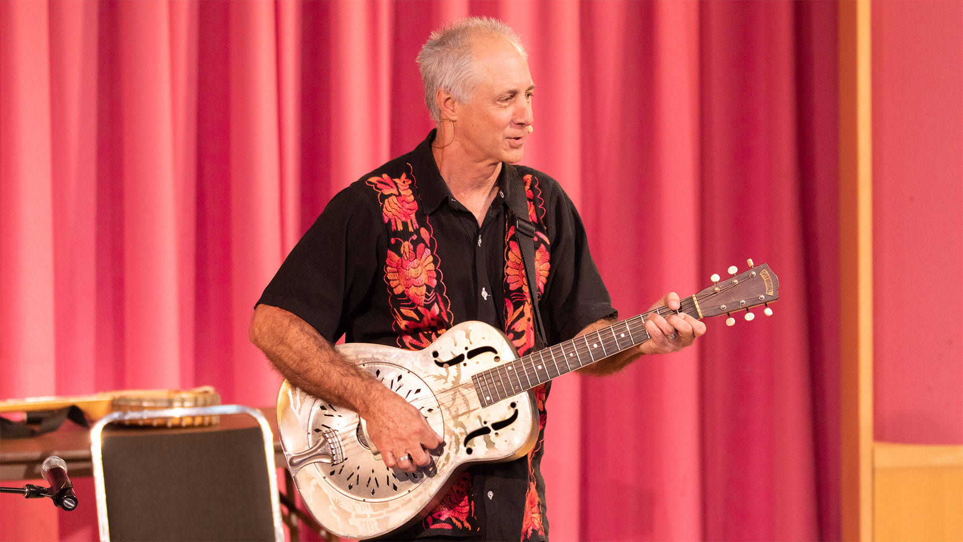 Stuart Stotts performing on stage. He is an older caucasian man with very short gray hair wearing a black Hawaiian shirt with red designed stripes down the front. He is playing a metal acoustic guitar against a pink red curtain on stage.