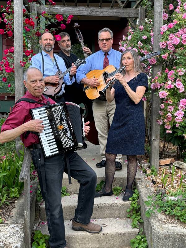 Five middle aged musicians stand outside on cement steps in front of pink flowers playing their instruments. 