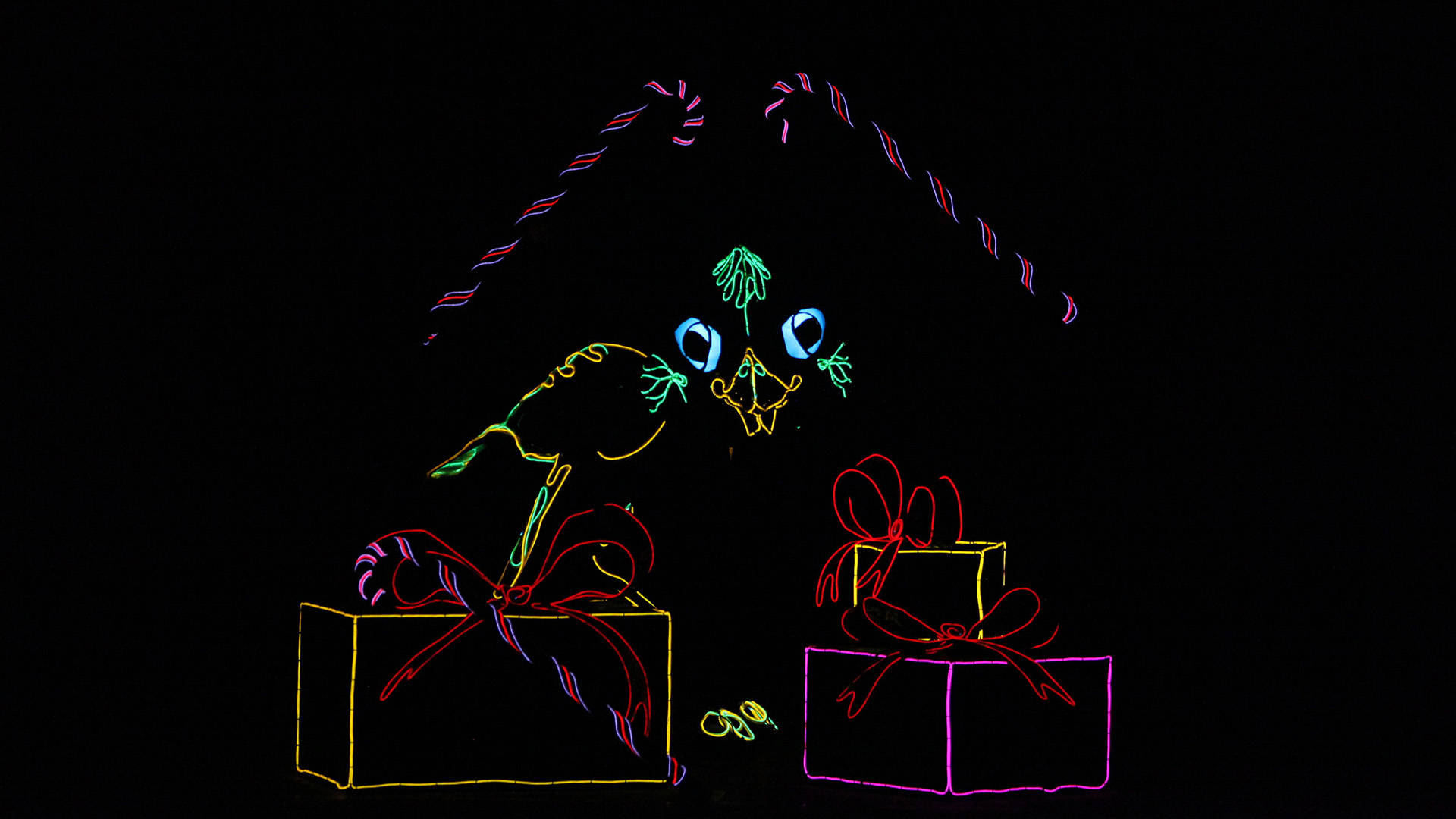 Neon light-up puppets and set pieces on an otherwise blackout stage. There is a bird, two candy canes and three presents with bows.