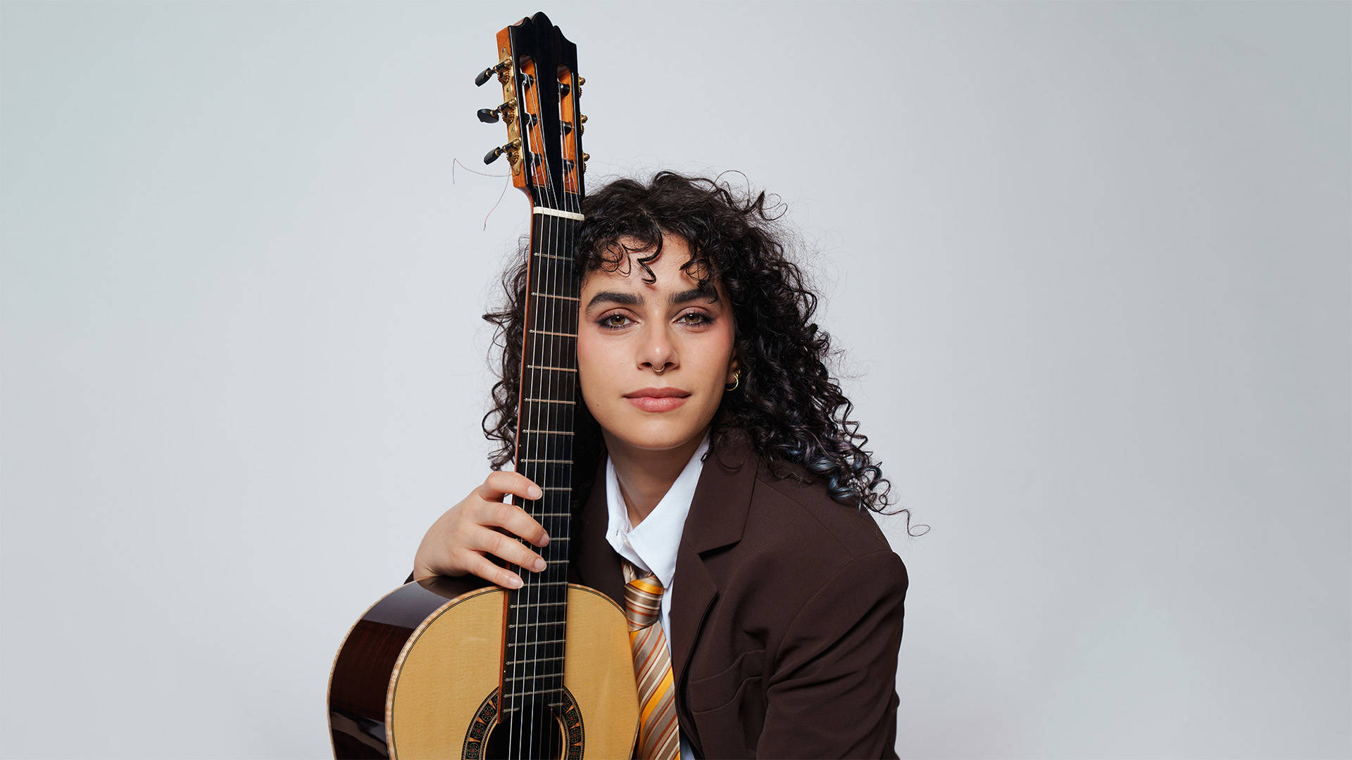 A woman with medium-light complexion, a septum piercing and long curly dark hair wearing a brown suit jacket, white shirt and yellow-brown tie, holding an acoustic guitar.