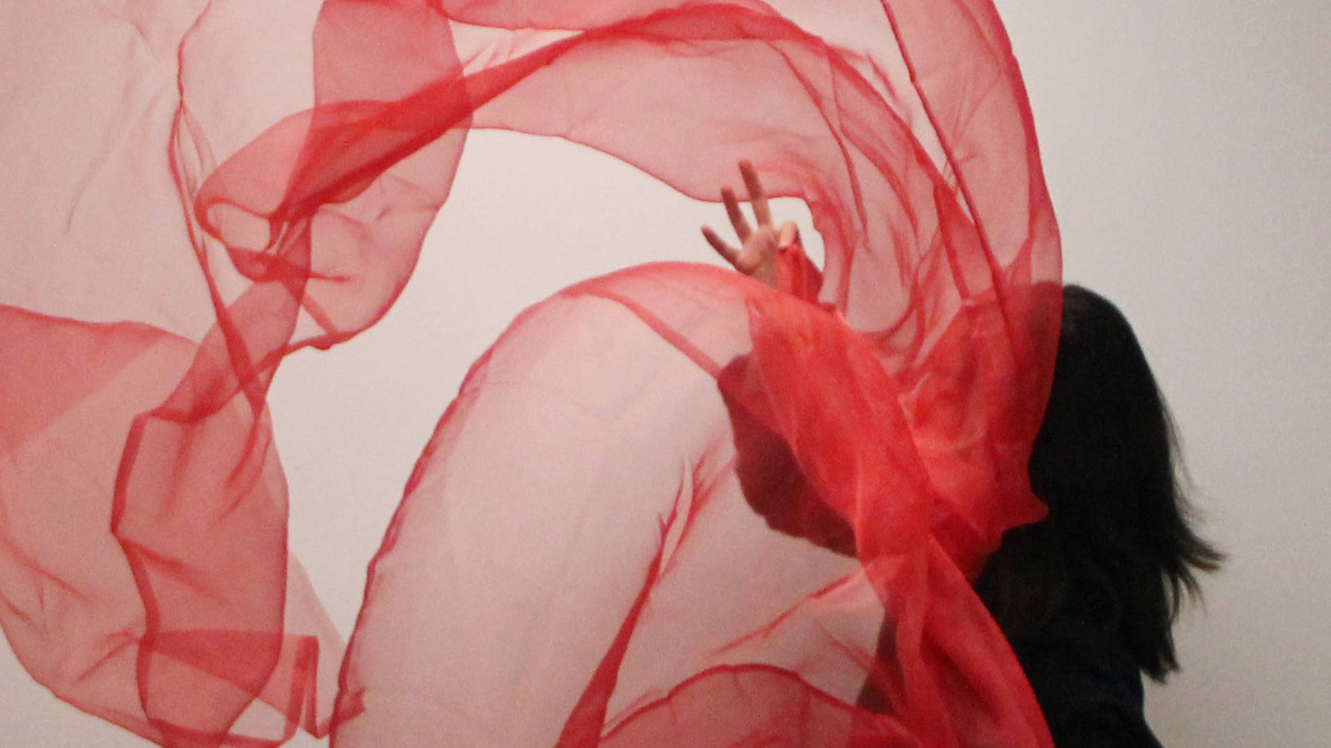 A woman with long black hair is throwing a long bolt of sheer red fabric against a white wall.
