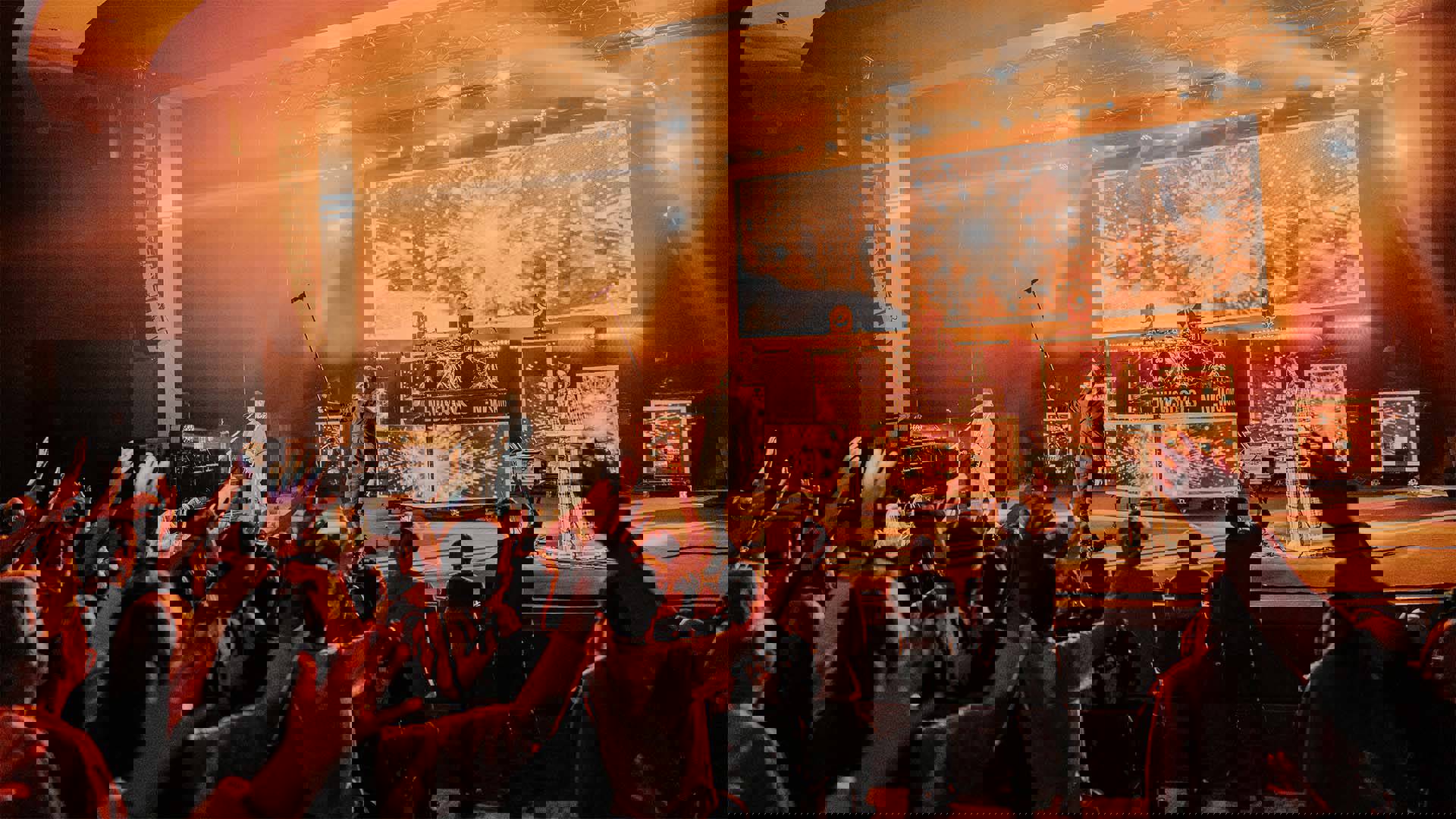 A rock band performing on a stage with an audience in the foreground with many hands raised in the air. The lighting is orange.