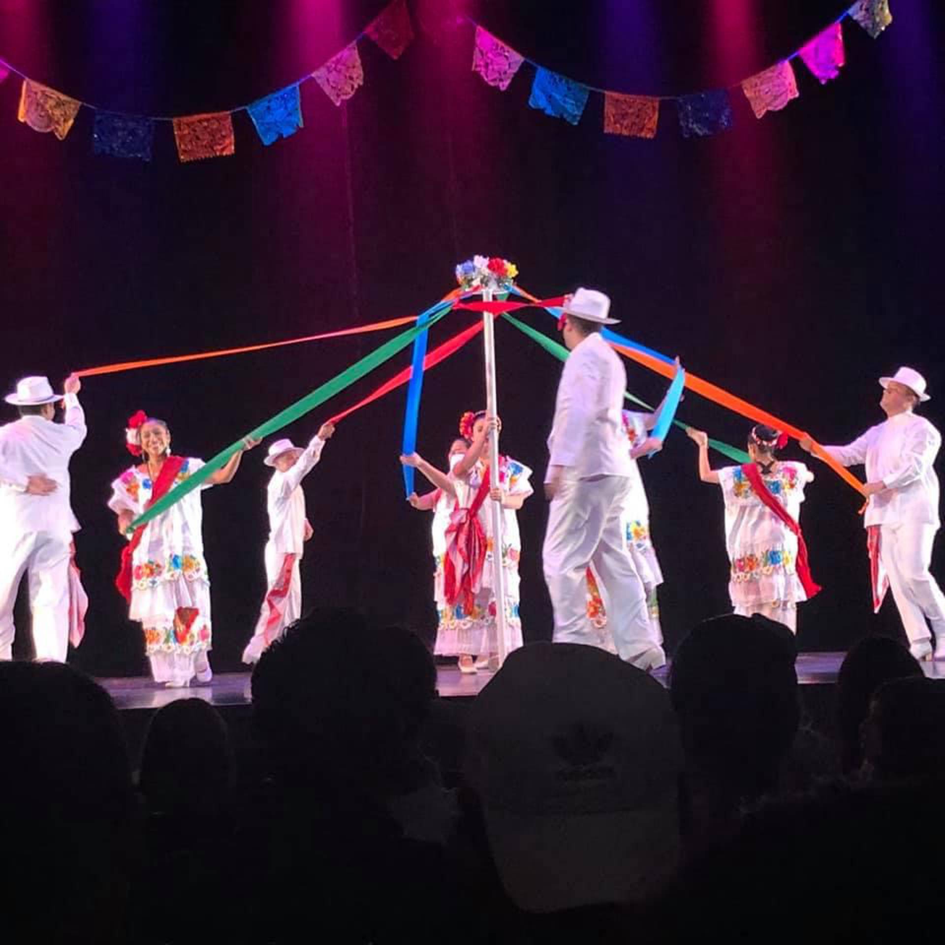 Dacners in white outfits with colorful floral patterns. They are standing around a pole, each holding a ribbon of a different color. The ribbons are attached to the top of the pole.