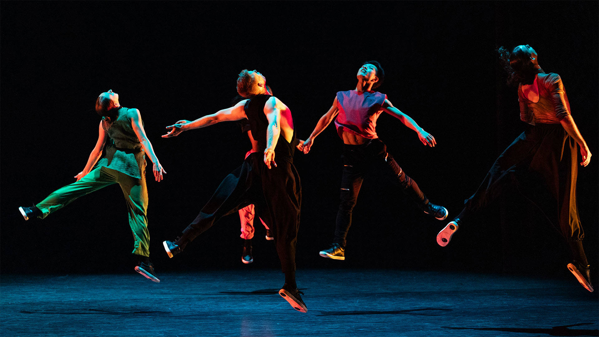 Four dancers in tank tops and loose pants mid-air with their arms out wide behind them, their legs out and the heads facing upwards. Stage lighting is dark with colored light reflecting on the dancers.