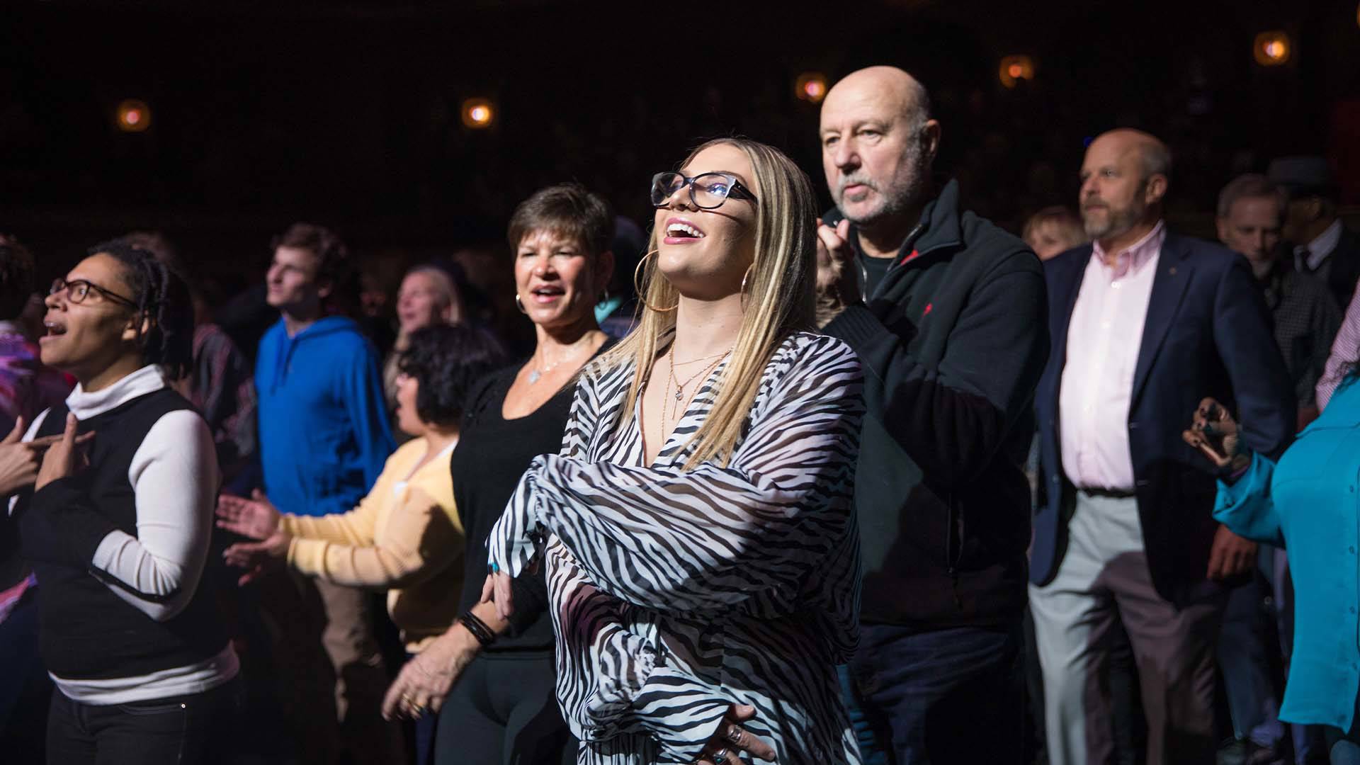 A group of people smiling and dancing looking up towards the bright lights of a stage.