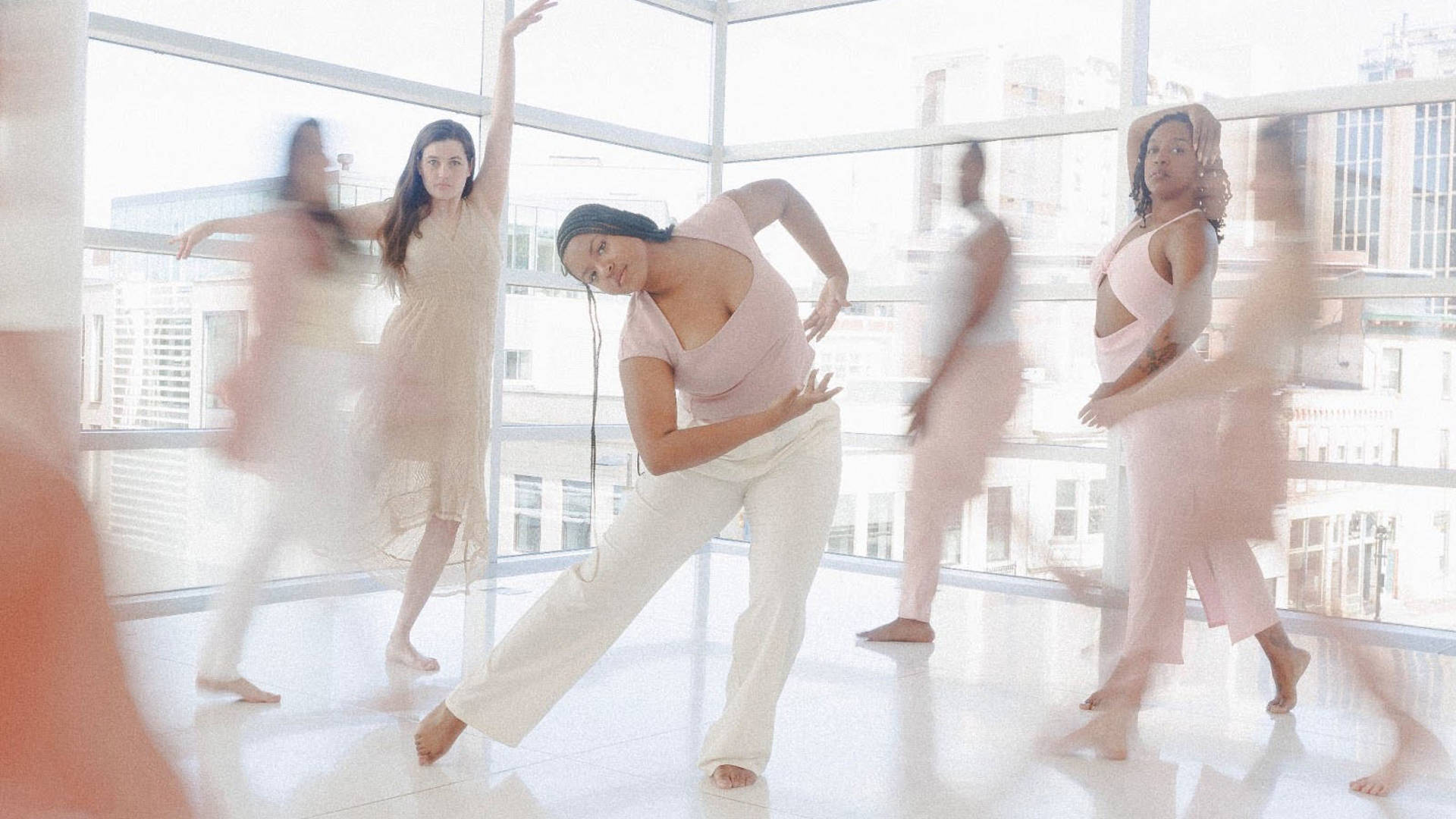 Dancers in various flowing light pink and white outfits striking dramatic dance poses against large windows looking out on buildings in the daytime.