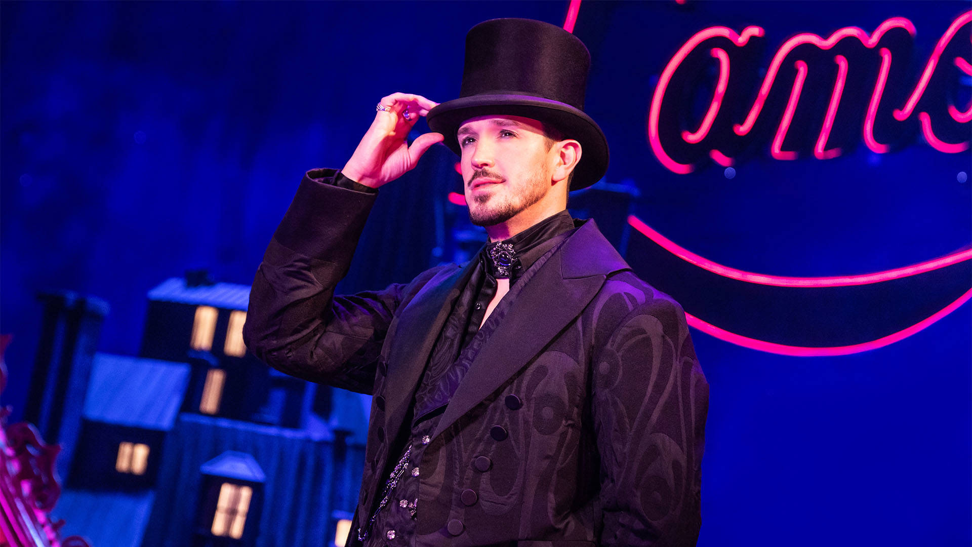 A caucasian man in a black silk suit with a black top hat. One hand is holding the hat's brim as he stares out off stage.