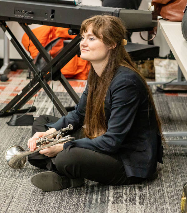 A young woman with light complexion and long brown hair sits on a carpeted floor holding a silver trumpet.