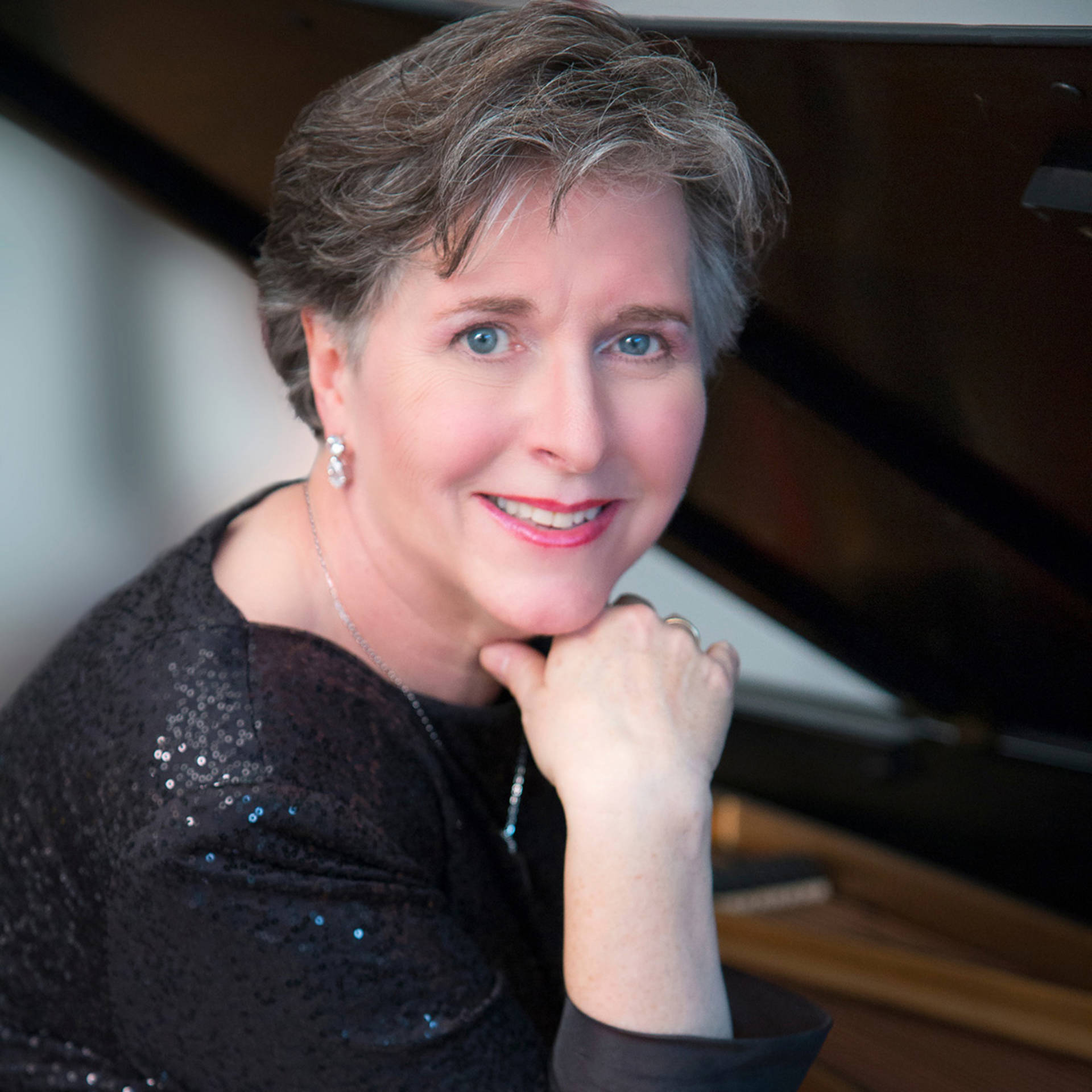 Headshot of an older caucasian woman with blue eyes and short greyish brown hair in a dark top with sequence. A blurry piano is behind her.