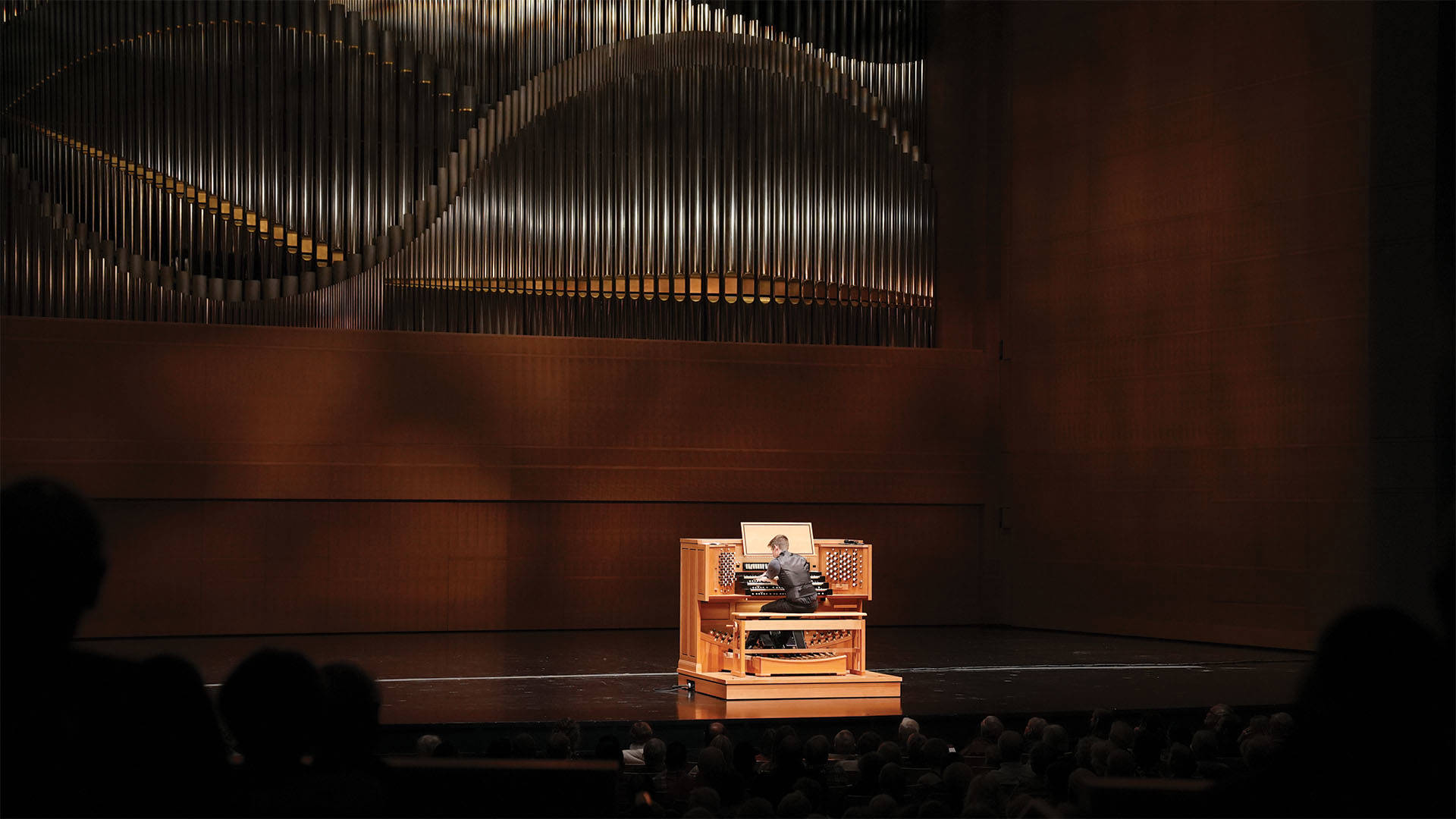 Greg Zelek, a young man with light complexion, short styled brown hair, a black button-up shirt and black vest, playing a large wooden organ console on a large stage with giant metalic pipes in the background.